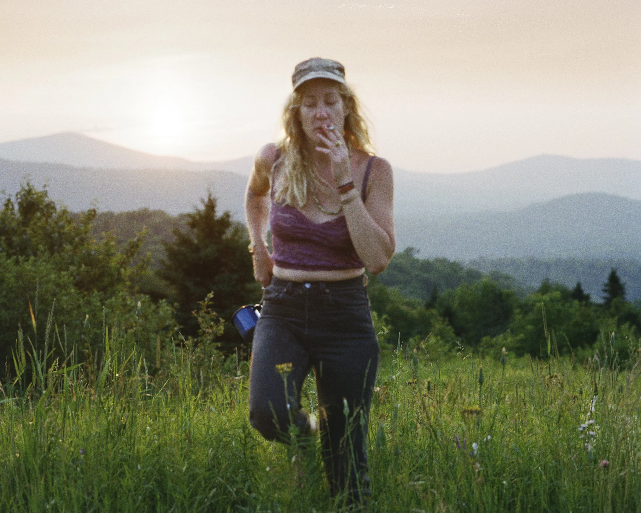 A young woman running through a grassy field at sunset, wearing a purple crop top, black pants, a cap, and smoking a cigarette, with mountains and forest in the background.