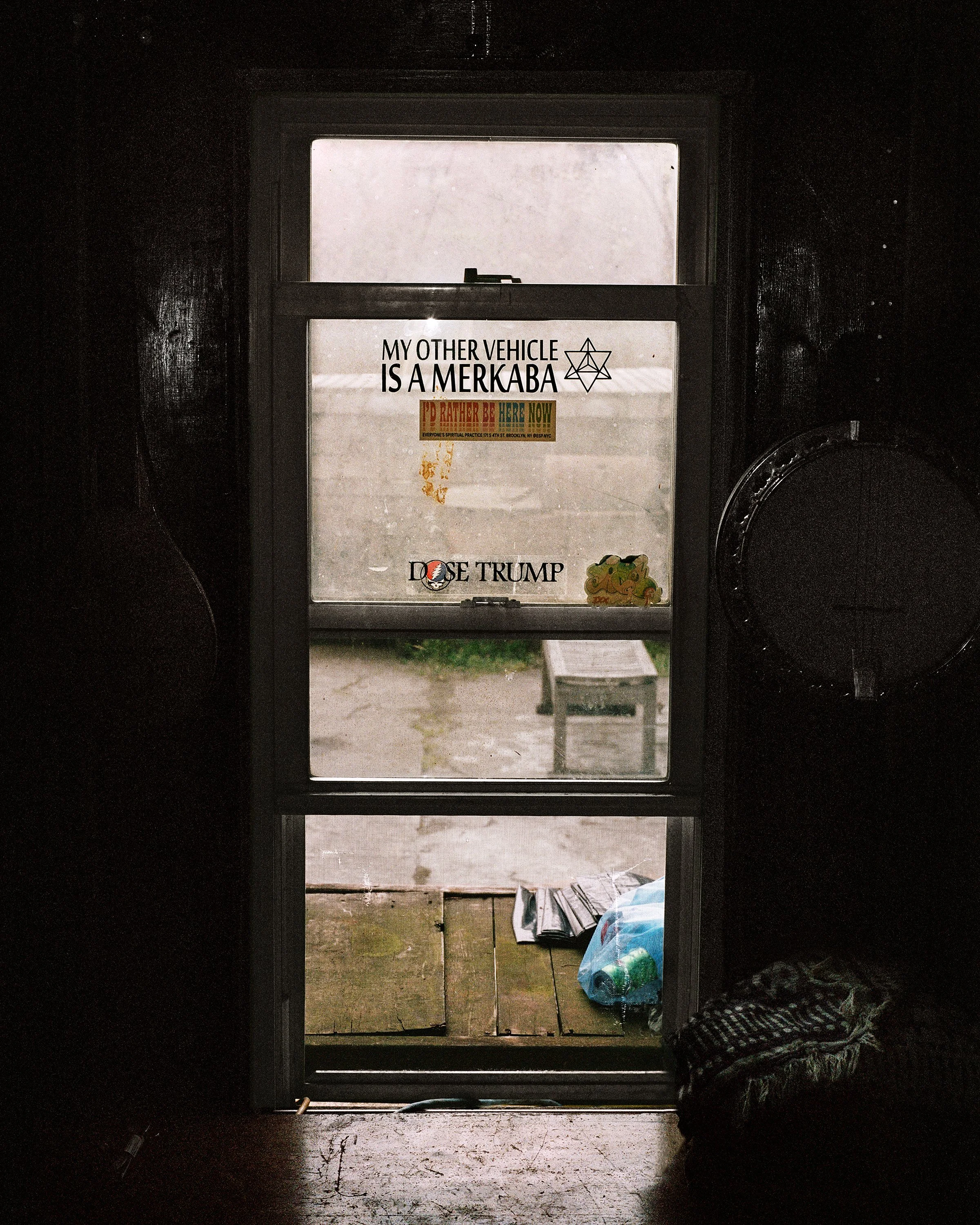 View through a glass door with multiple stickers, including one that says "My other vehicle is a mercaba," another with a rainbow-colored message, and a sticker that says "Dose Trump." Outside the door, there are a wooden porch and some grass, with a