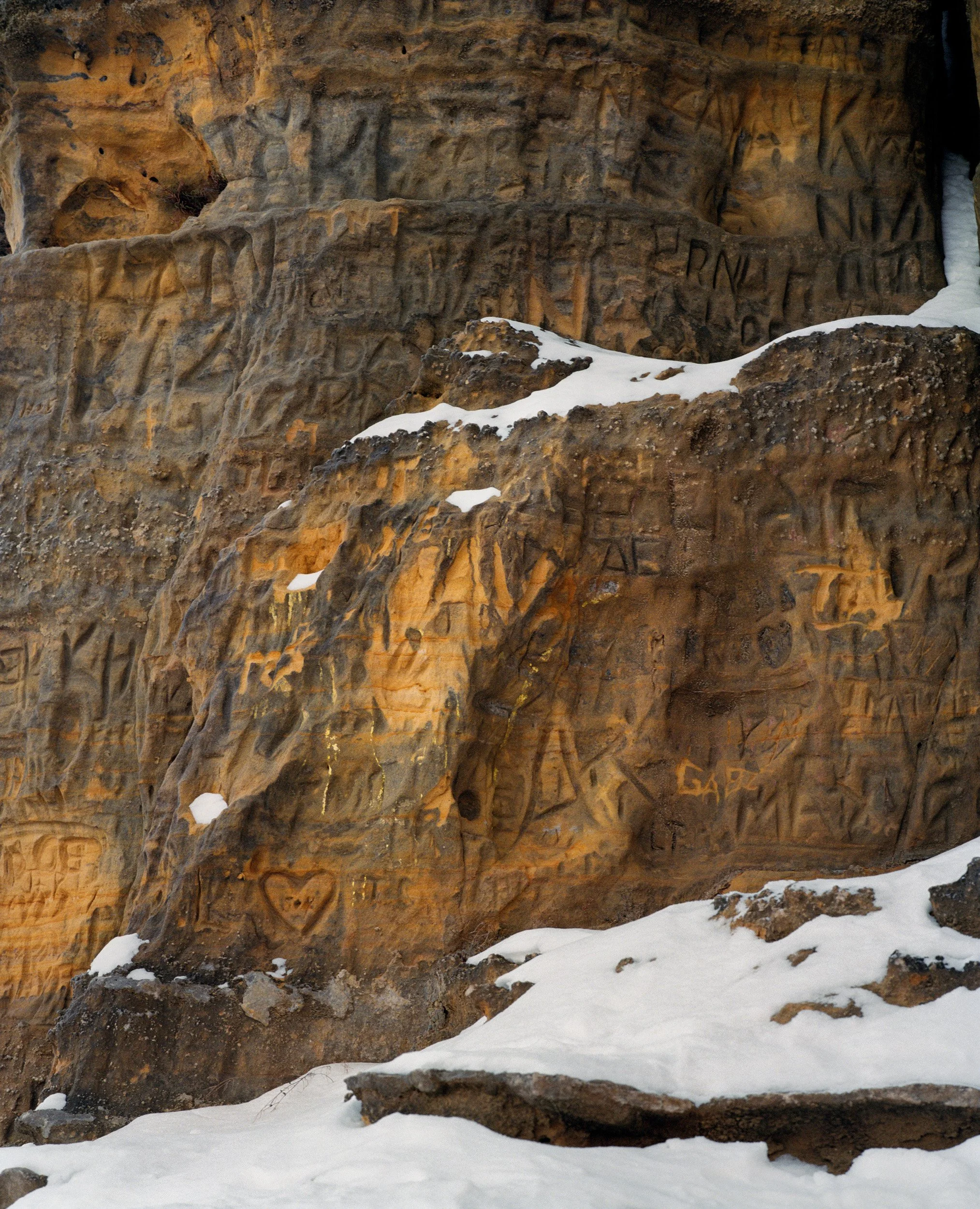 Rock face with numerous carvings and graffiti, partially covered in snow.