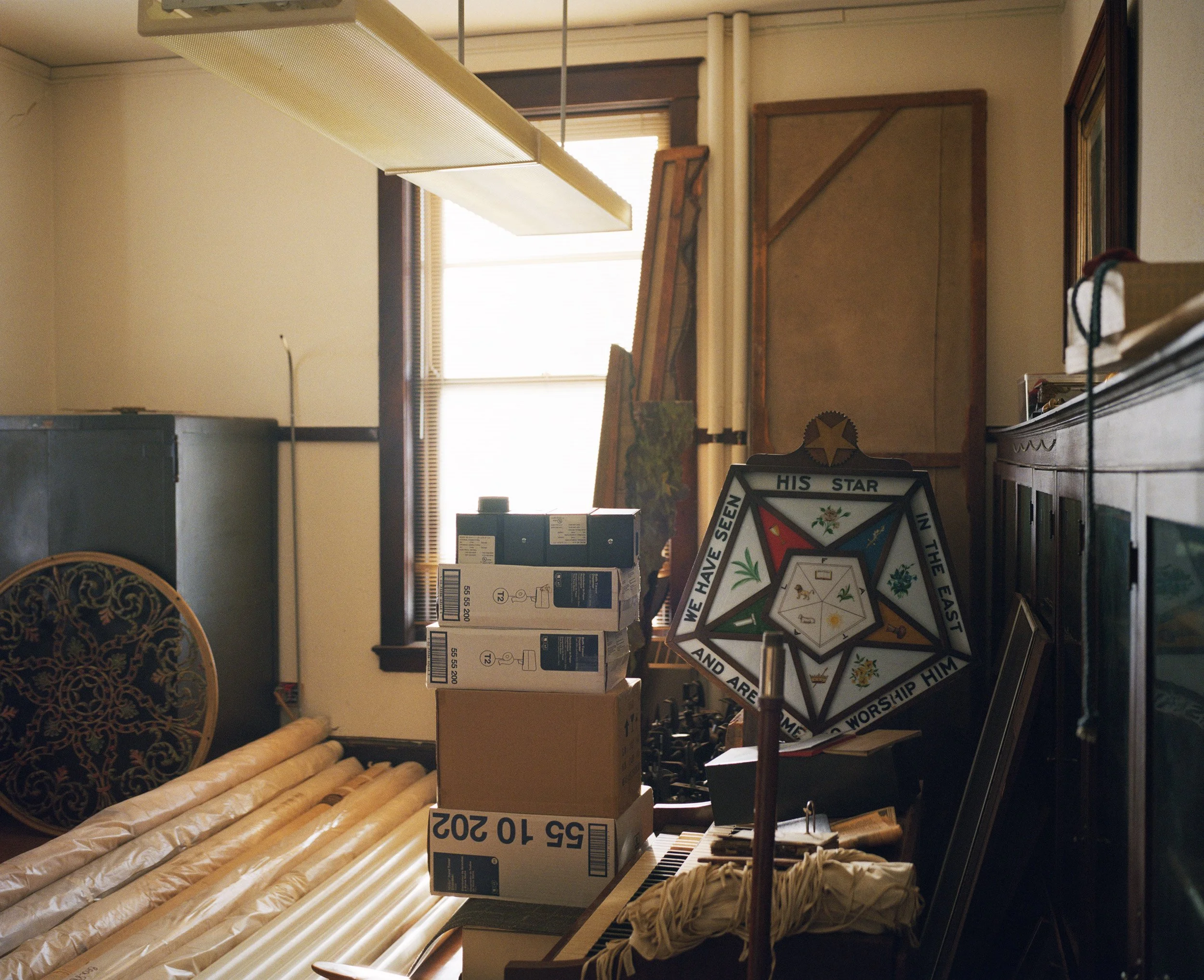 A cluttered room with boxes, artwork, and miscellaneous items. A stained-glass decoration with religious text and symbols is visible near a window.