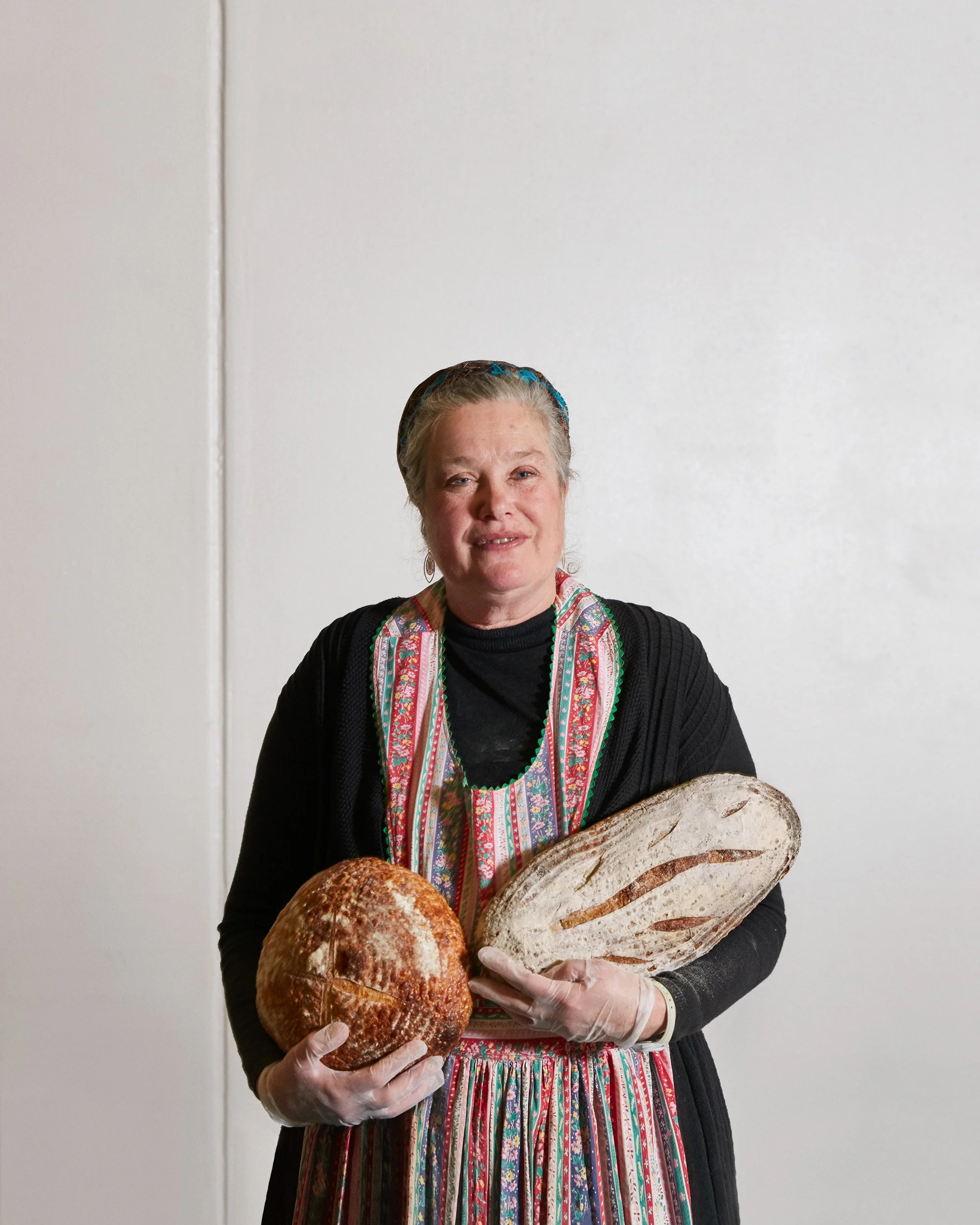 An older woman with a headscarf holding two loaves of bread, standing against a plain white wall.