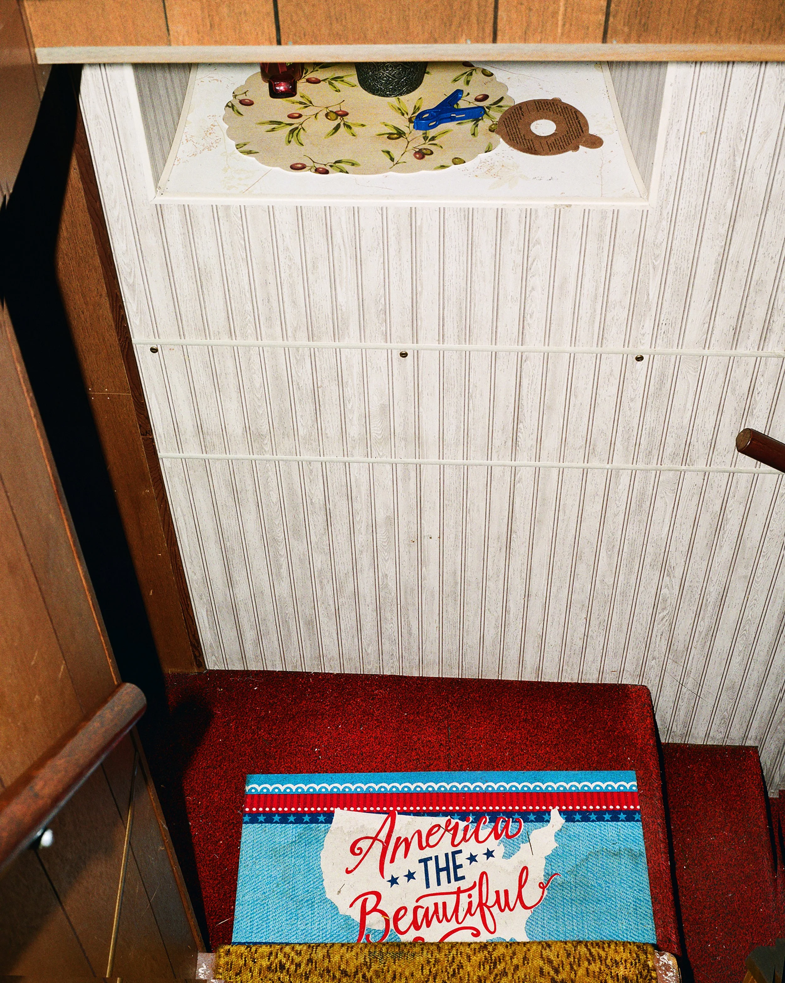 Photo of a countertop with a decorative placemat, a small potted plant, a blue clothespin, and a round coaster. Below is a red carpeted staircase with a patriotic rug that has an outline of the United States and the words "America the Beautiful."