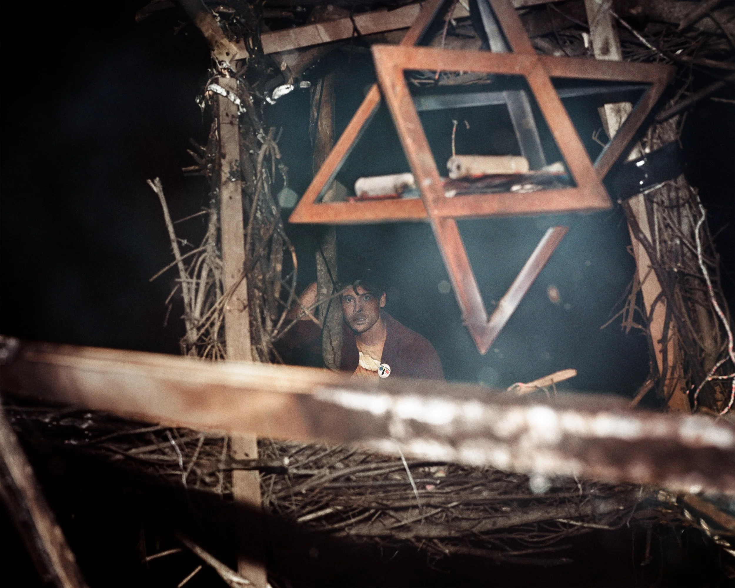 A man with dark hair and a serious expression is looking up from a dark and chaotic environment filled with wooden sticks and debris. A large wooden Star of David hangs overhead, partially obscured by the clutter.