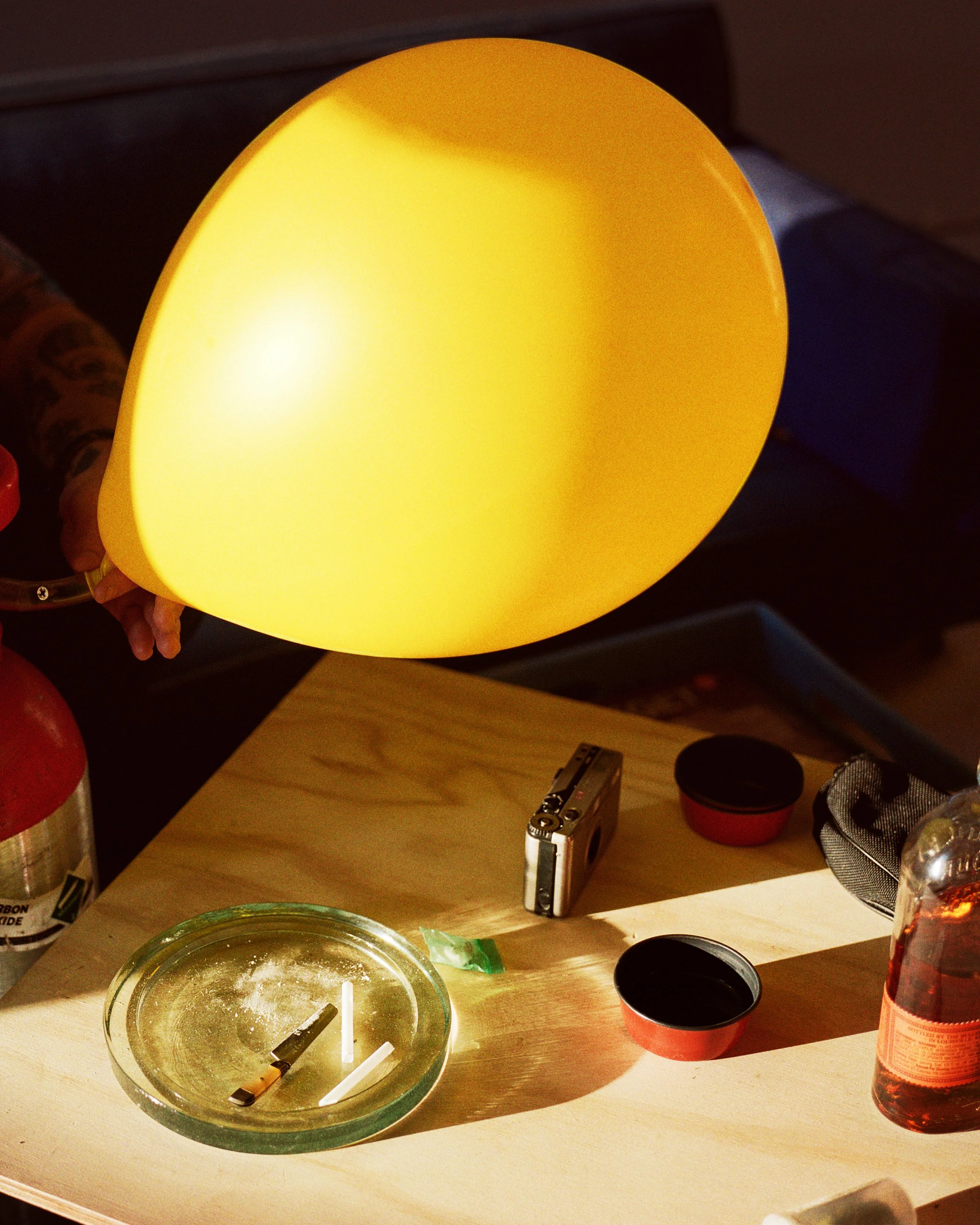 A yellow balloon floating above a wooden table with various objects including a cigarette, an ashtray, a disposable camera, and small containers.