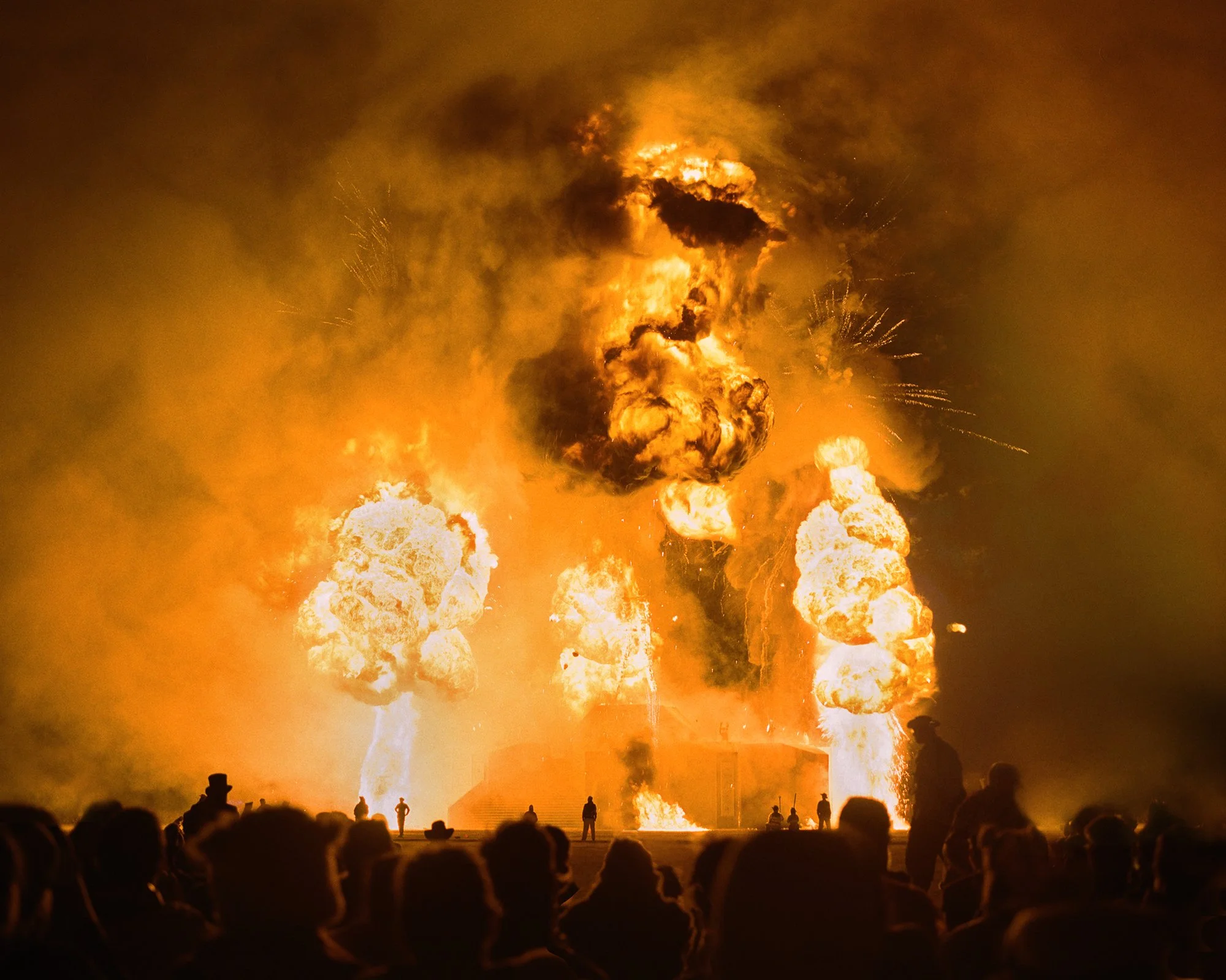A large explosion with fire and smoke during a fireworks display at night, with silhouetted spectators watching.
