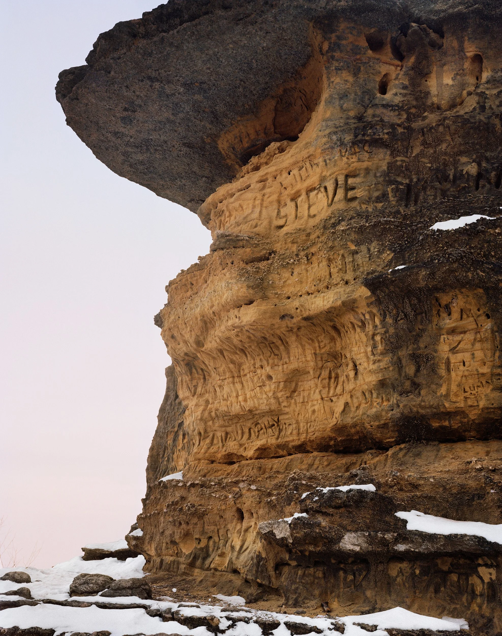 Close-up of a rock formation with carved engravings, some partially visible, in winter with snow at the base and a pale sky background.