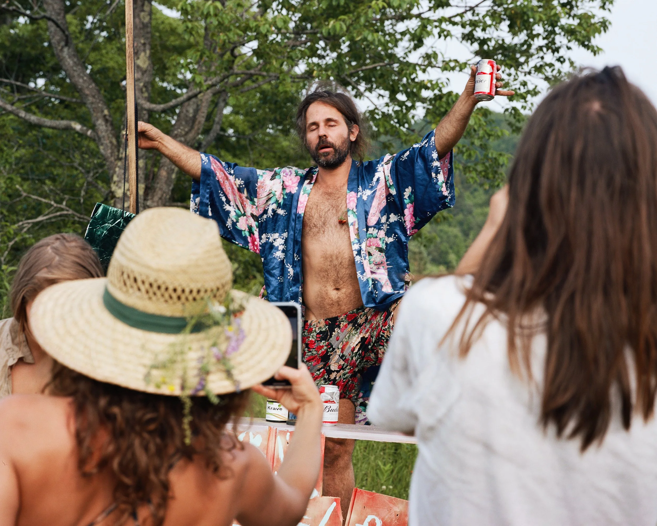 A man with long hair and a beard wearing a floral kimono with his shirt off, holding a can of beer in one hand and raising his arms, standing outdoors surrounded by trees. Several women are in front of him, one wearing a large straw hat and taking a
