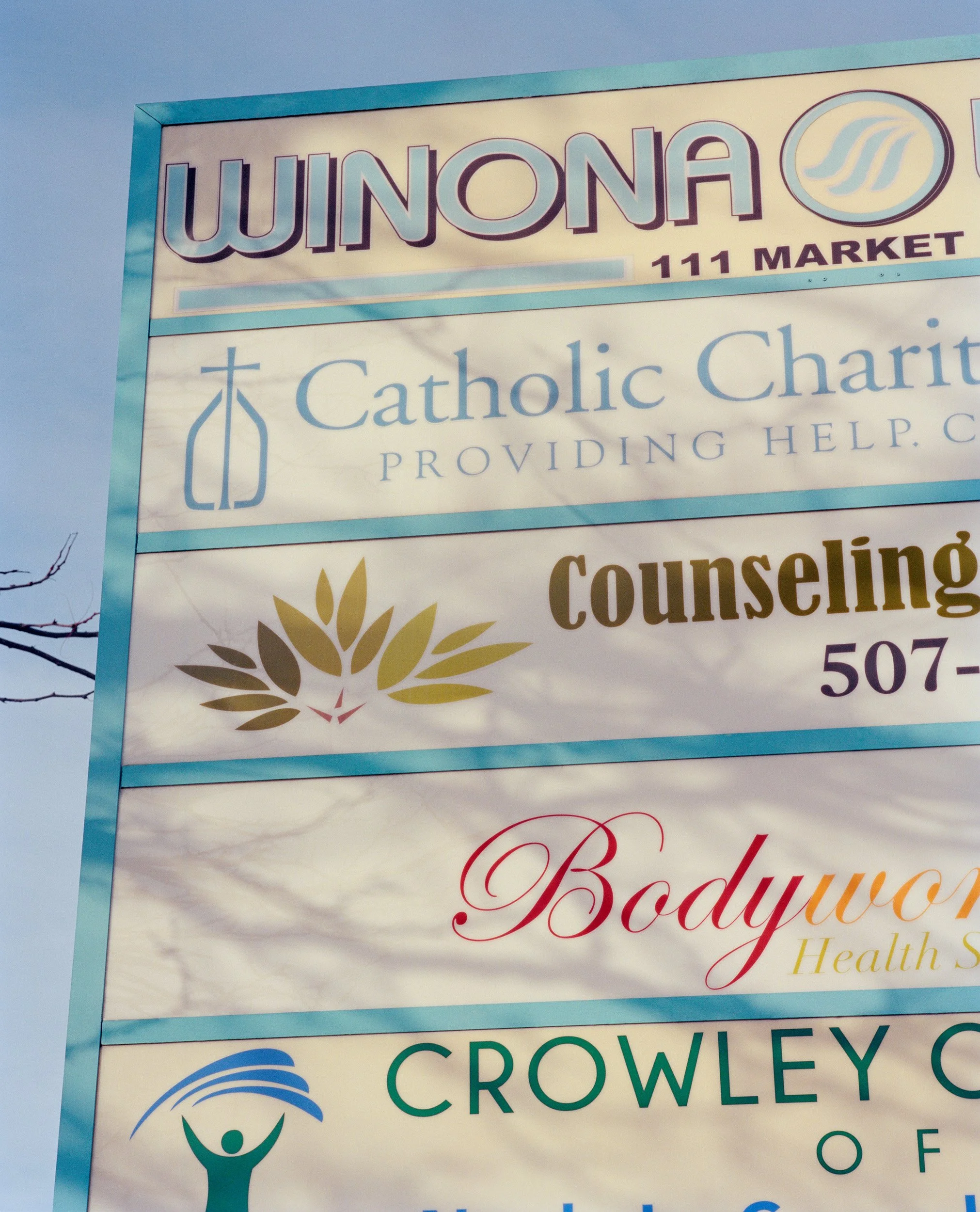Outdoor sign with multiple business advertisements, including WINONA 111 MARKET, Catholic Charities, Counseling, Bodyworks Health, and Crowley.