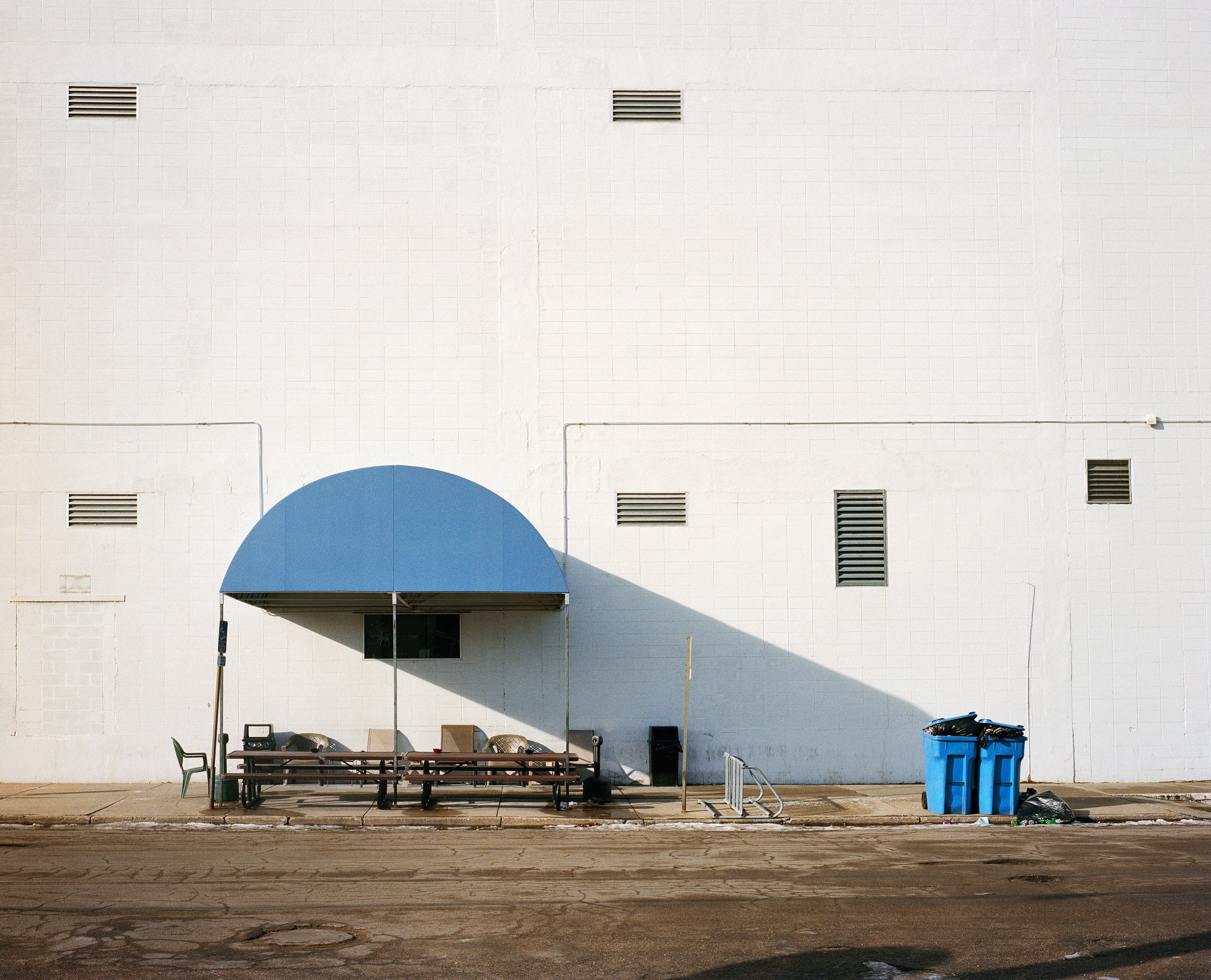 Empty bus stop with benches under a blue awning, surrounded by trash bins and a white wall with small vents.