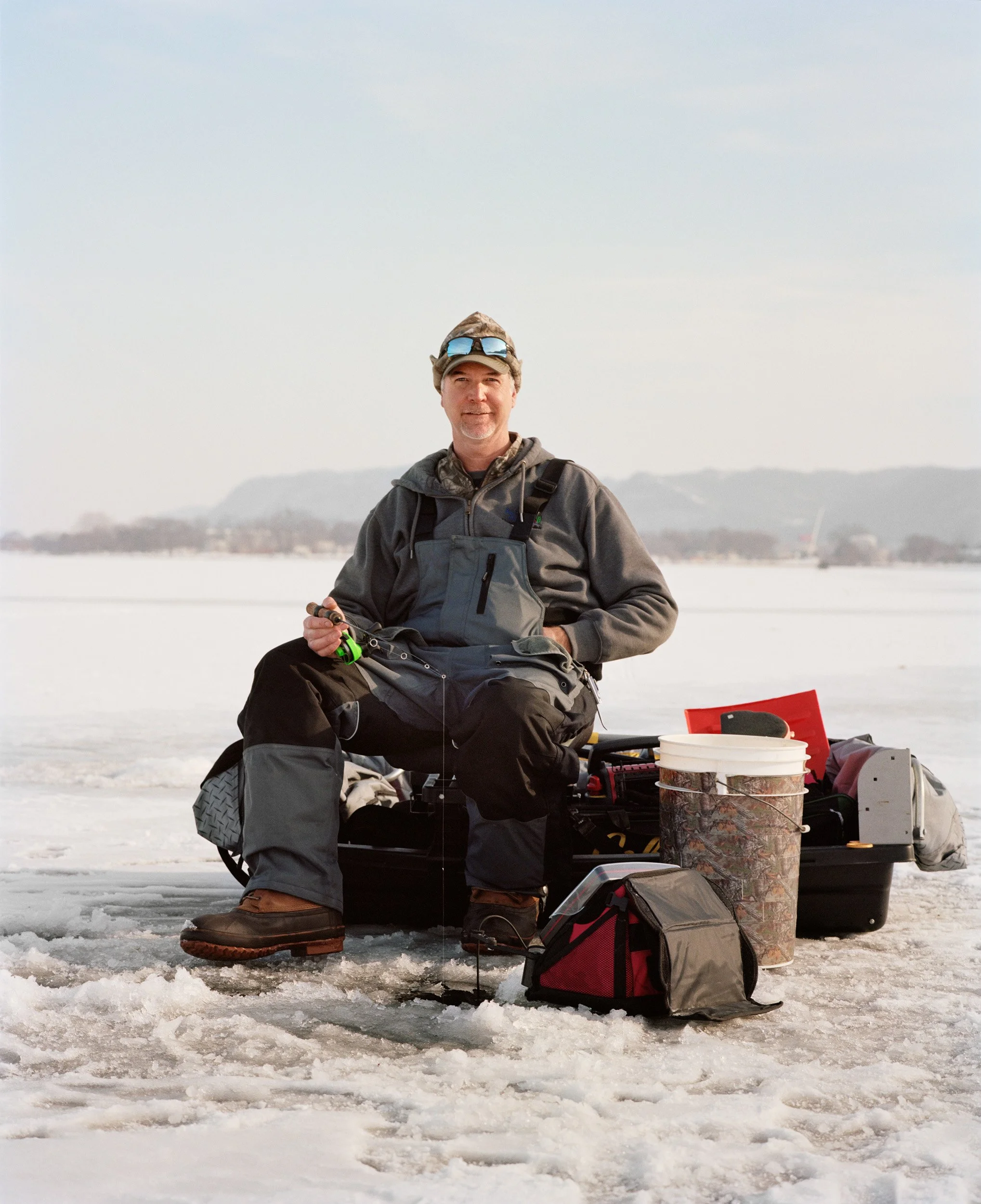 A man ice fishing on a frozen lake, sitting on a small folding chair surrounded by fishing gear, buckets, and supplies.