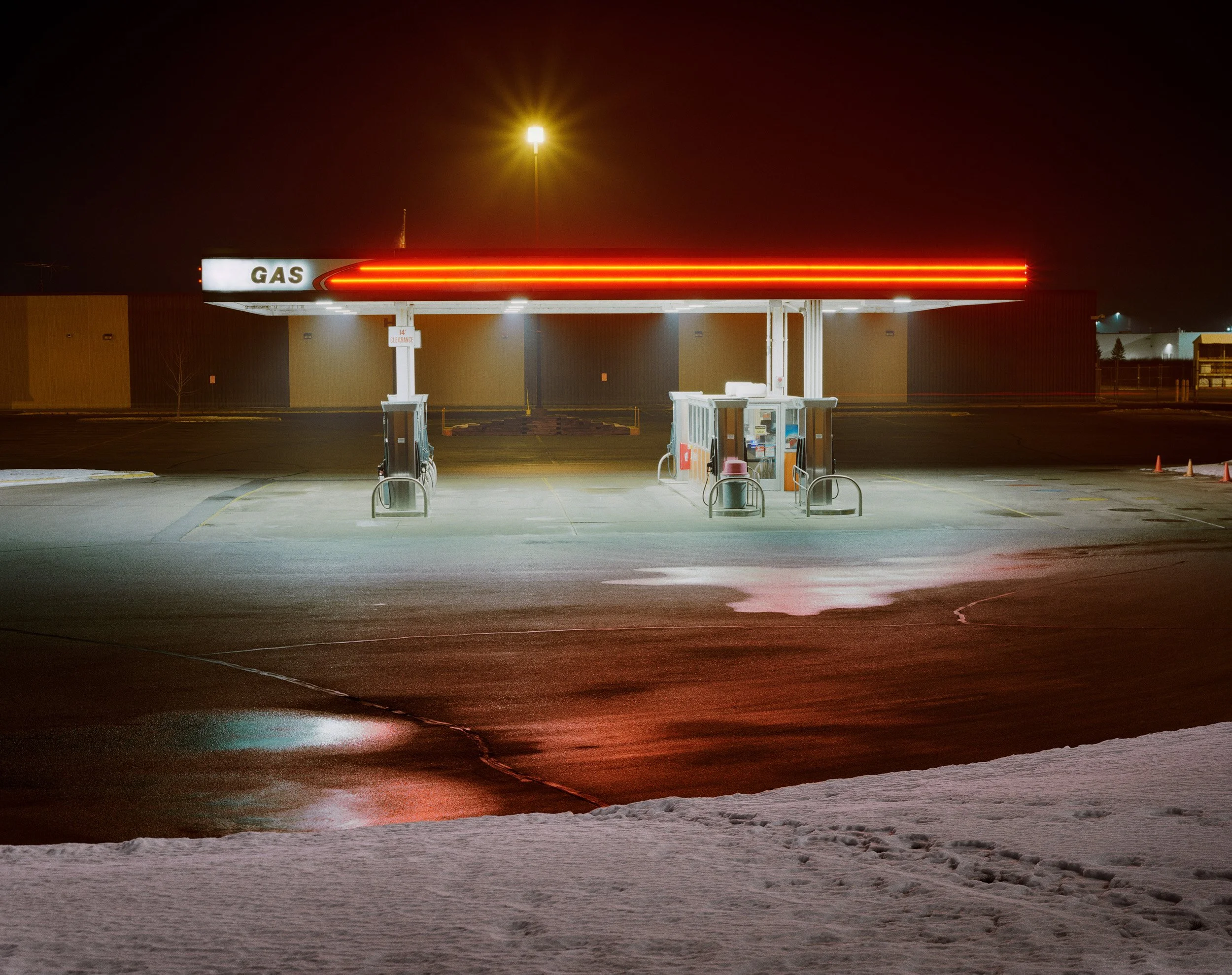 Empty gas station at night with illuminated signs and wet pavement, snow on the ground.