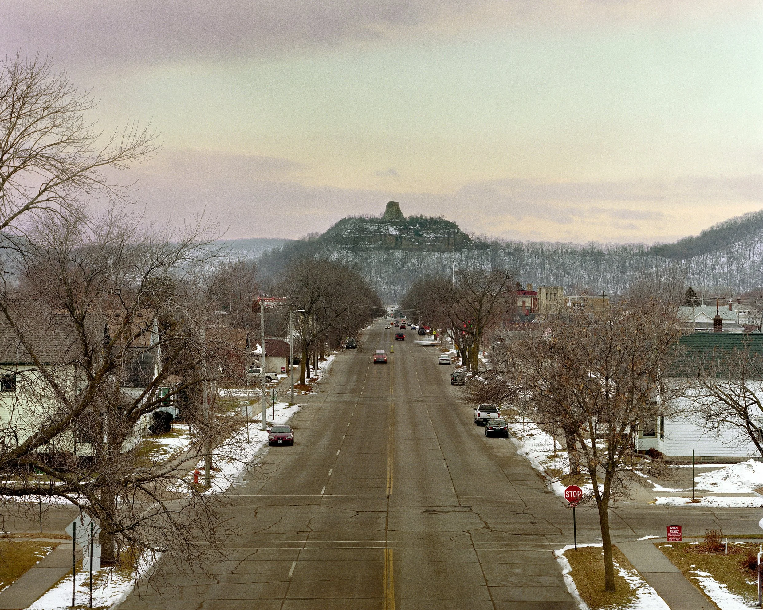 An empty street in a small town during winter, with snow on the ground and bare trees on both sides. In the distance, a hill with a distinct rock formation on top is visible under a cloudy sky.
