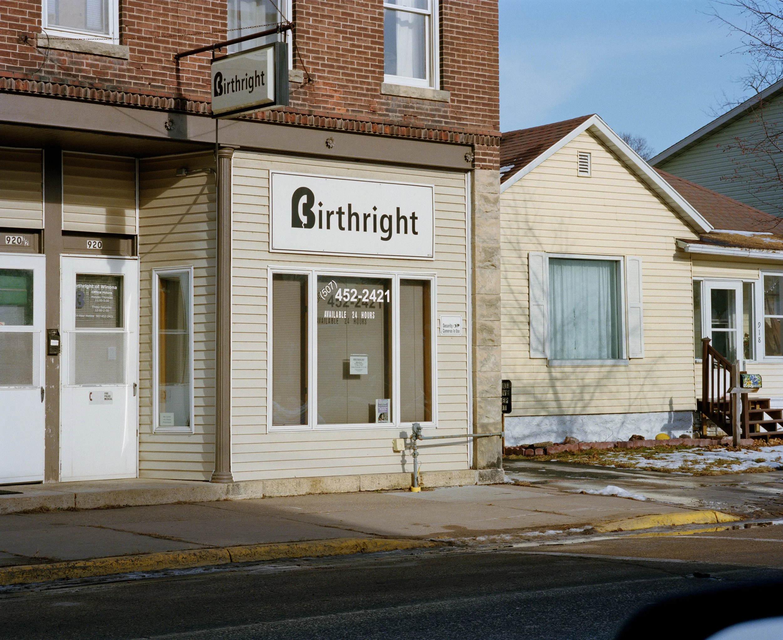 A small commercial building with a sign reading 'Birthright' with a phone number in the window, next to a yellow house with a small porch, in a town on a winter day.