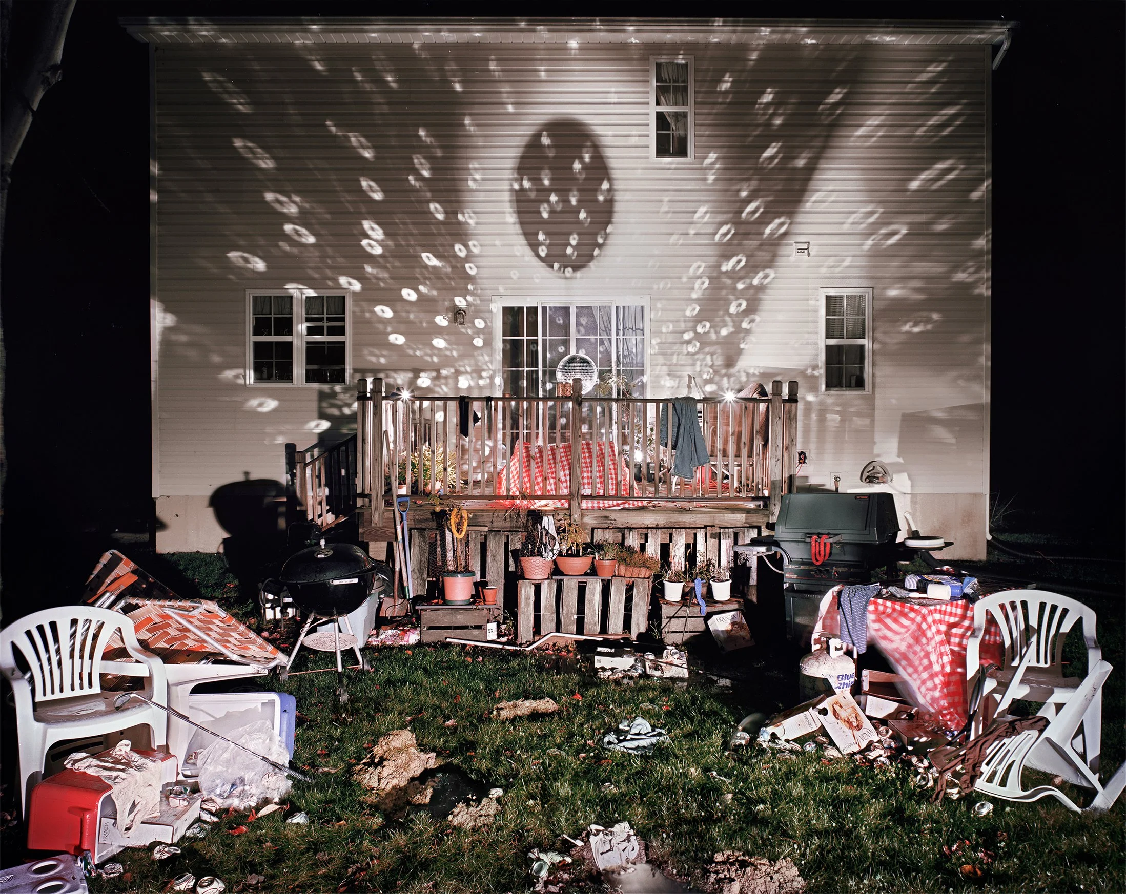 Nighttime exterior view of a house with a porch lit by string lights, casting a pattern of light and shadow on the house's wall. The yard is cluttered with overturned lawn chairs, a barbecue grill, and various items, indicating recent activity or a s