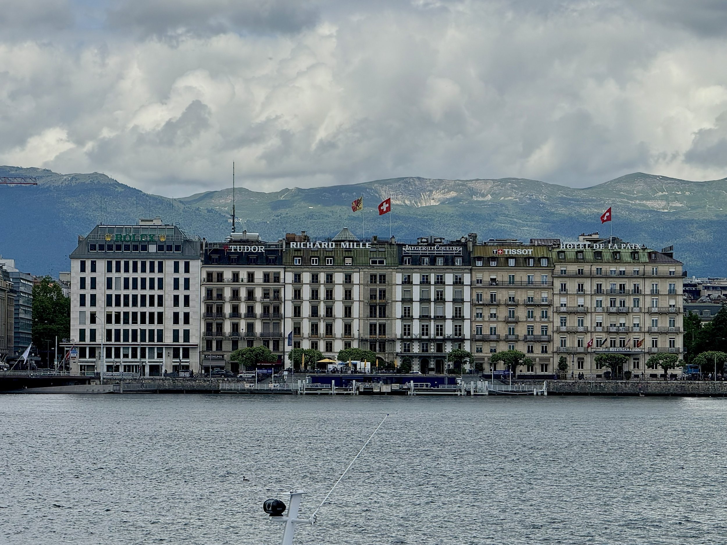 A cityscape with multiple buildings along the water, with mountains and cloudy sky in the background. Flags, including the Swiss flag, are on top of the buildings. The water has a boat and a fishing rod in it.