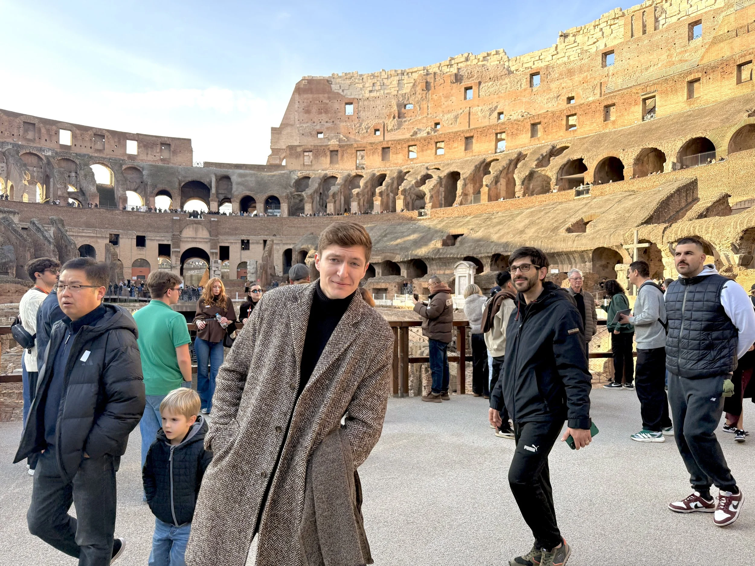 Tourists visiting the Colosseum in Rome, Italy during the daytime, with some looking around and taking photos, and a young man in the foreground wearing a checked coat.