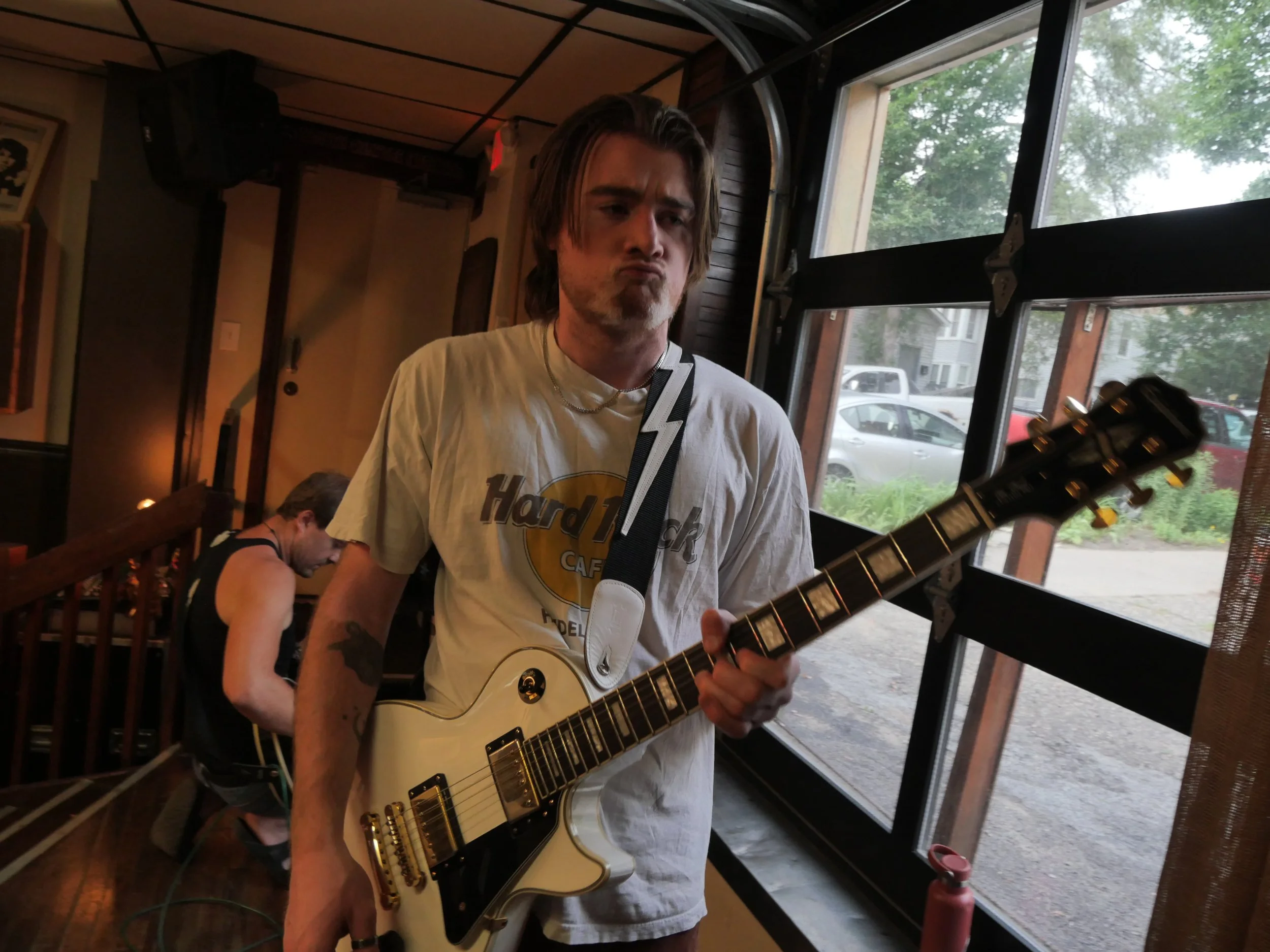 A young man with long hair playing a white electric guitar indoors near a large window with trees outside. There are other people in the background, one sitting at a table.