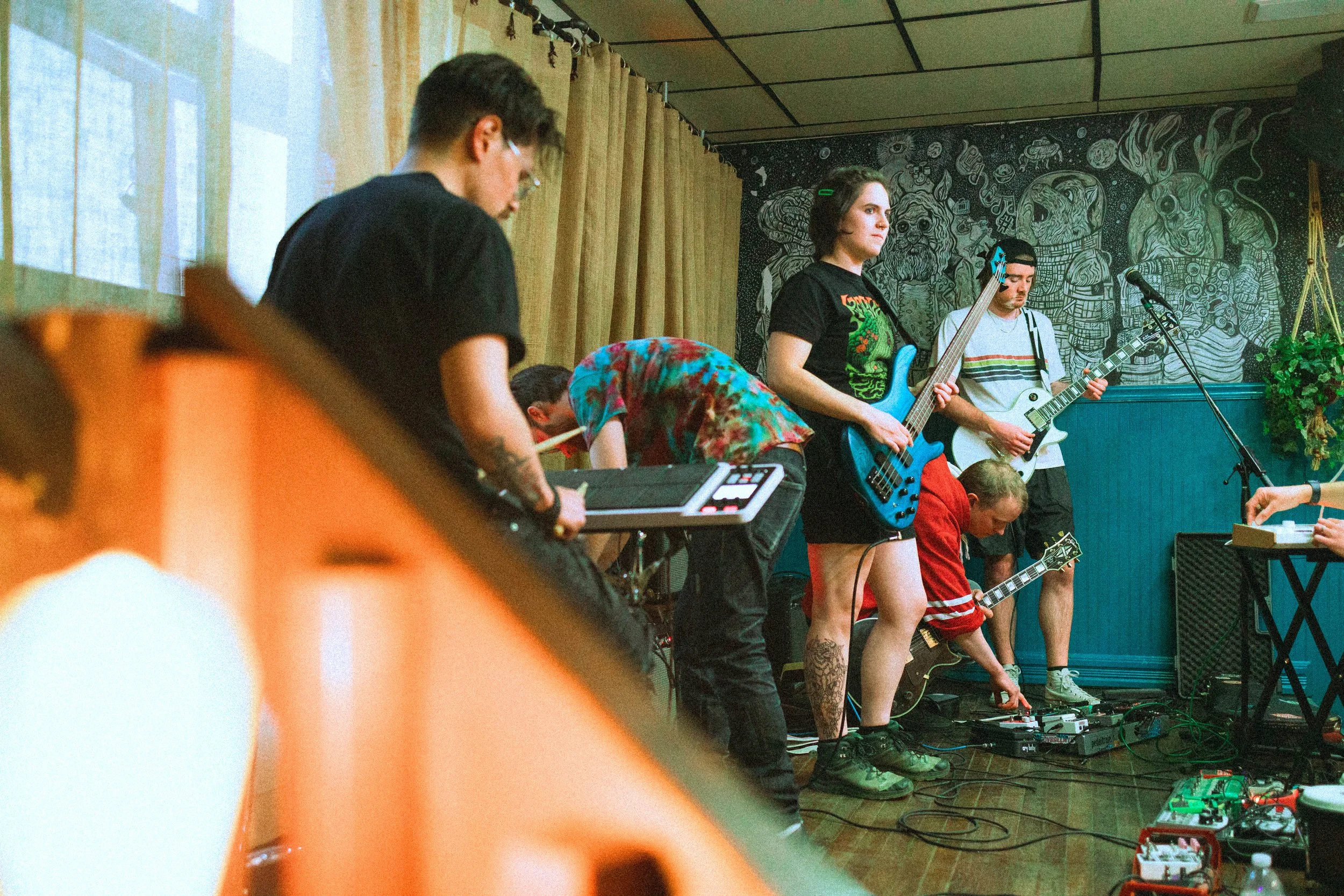 A group of musicians performing on stage, with some playing guitars and others using electronic equipment, in a room with a mural wall in the background.