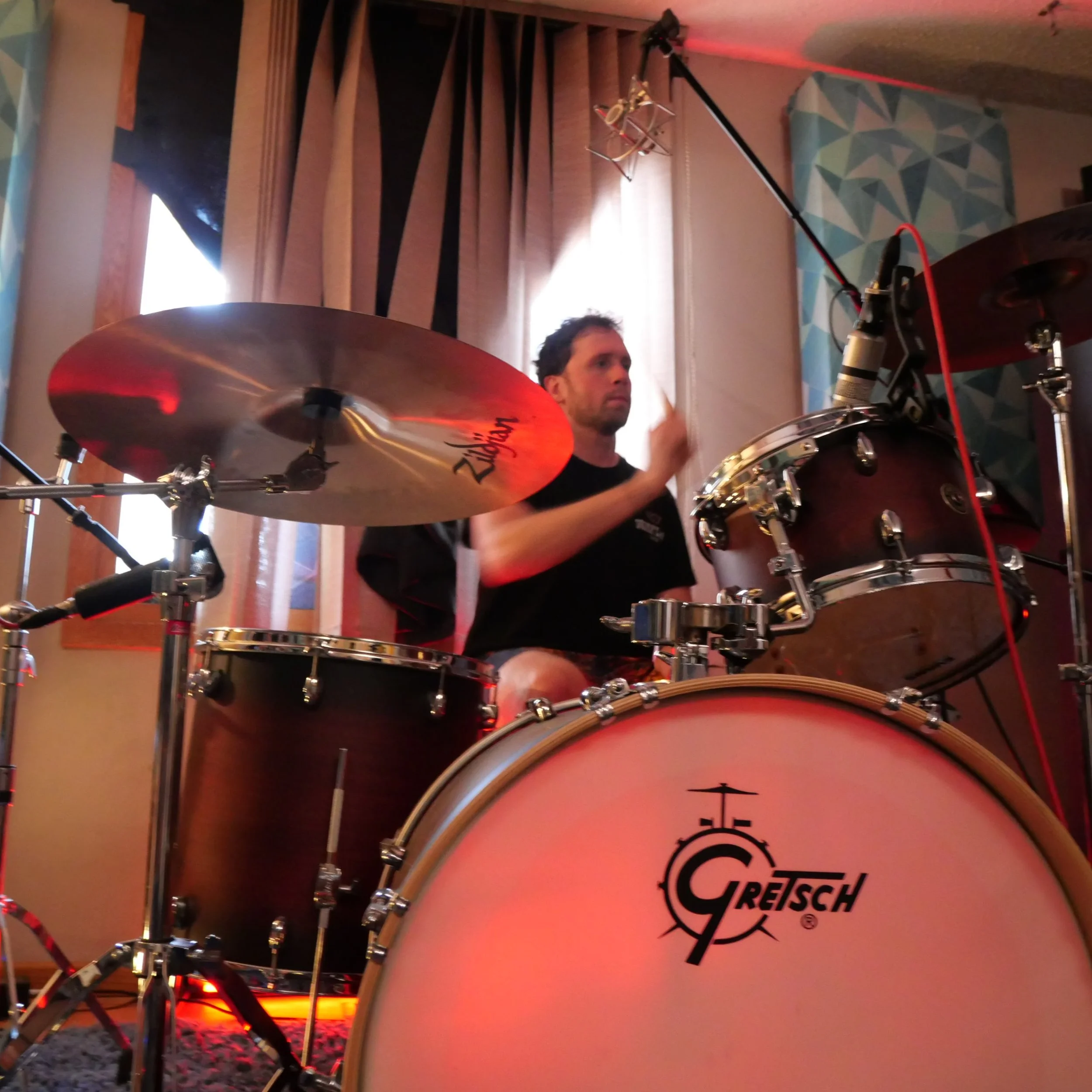 Close-up of a man playing a drum set with a crash cymbal in an indoor setting. The drums have a logo that says 'Gretsch.' The background features a window with wooden blinds and a wall with geometric patterned wallpaper, with red lighting accents.