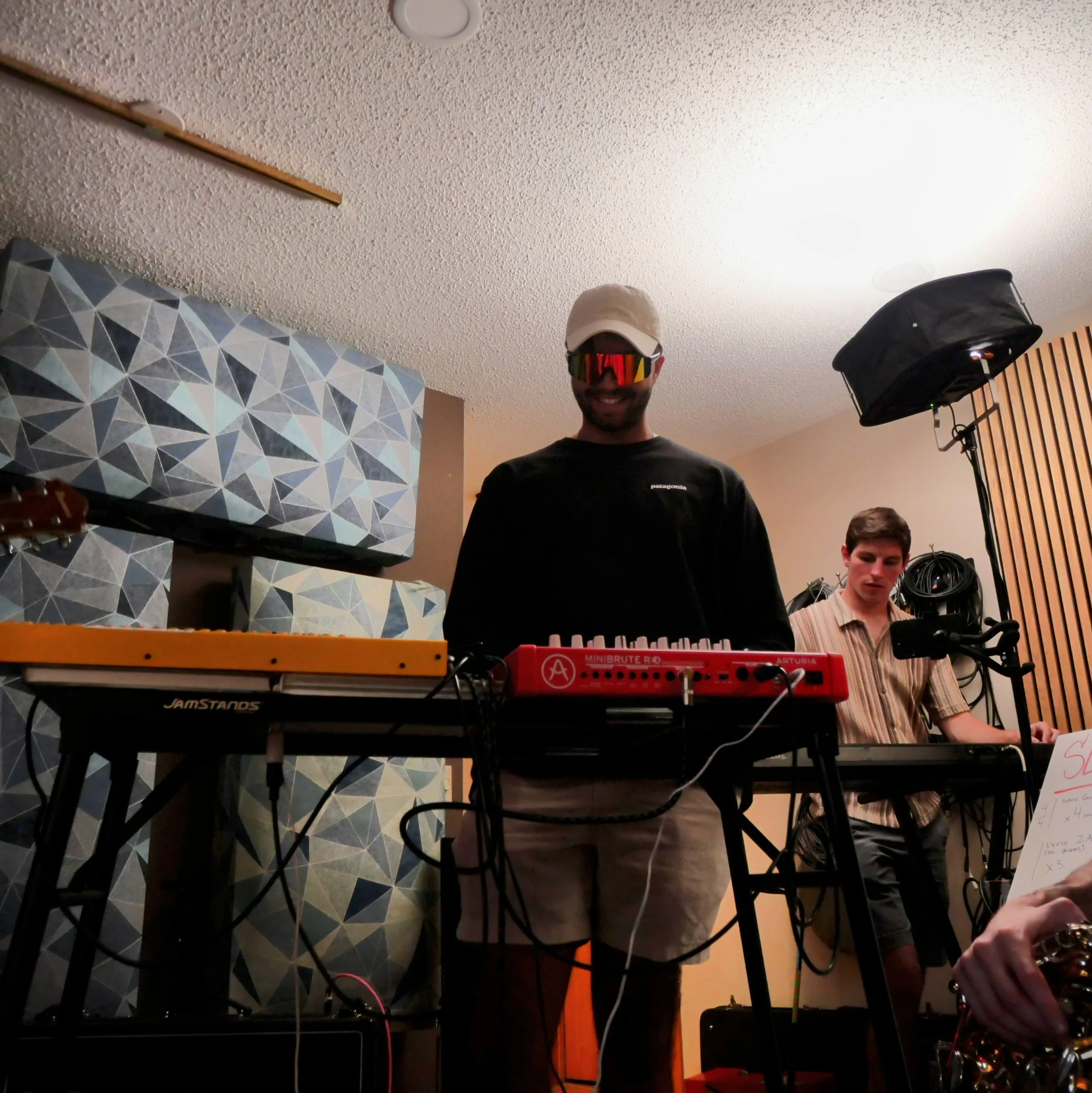 Photo of a smiling man wearing sunglasses, a beige cap, black shirt, standing in a music studio with a keyboard, equipment, and notes on a notebook in front of him.
