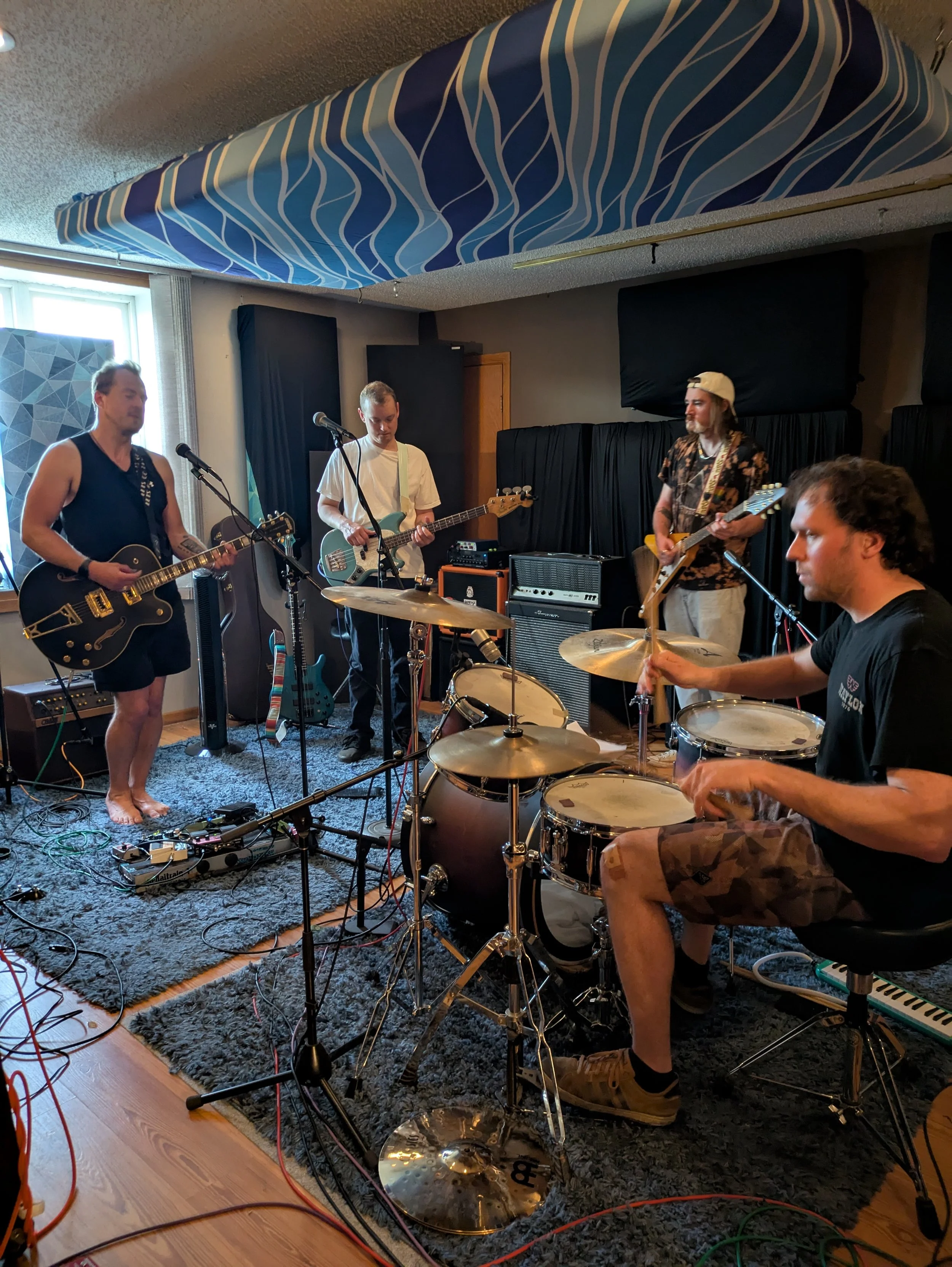 A band of four musicians performing indoors, with electric guitars and a drum set, in a room with a window, black curtains, sound equipment, and a blue patterned acoustic panel on the ceiling.