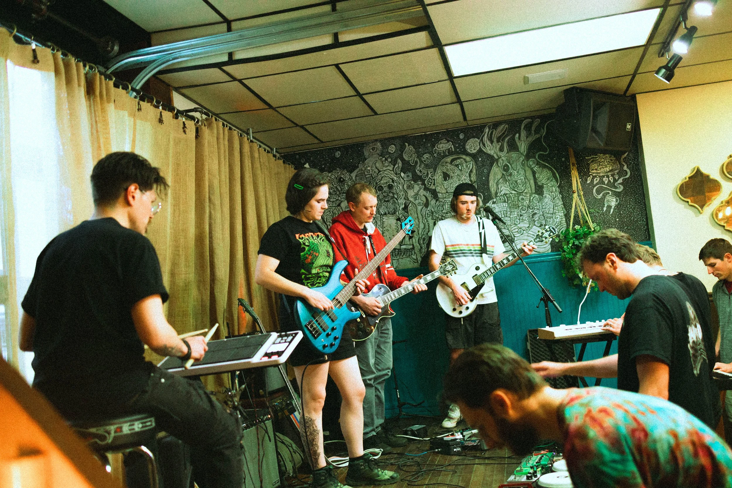 A group of young musicians performing on stage with guitars, keyboard, and electronic equipment, with a mural of space-themed artwork in the background and an audience watching.