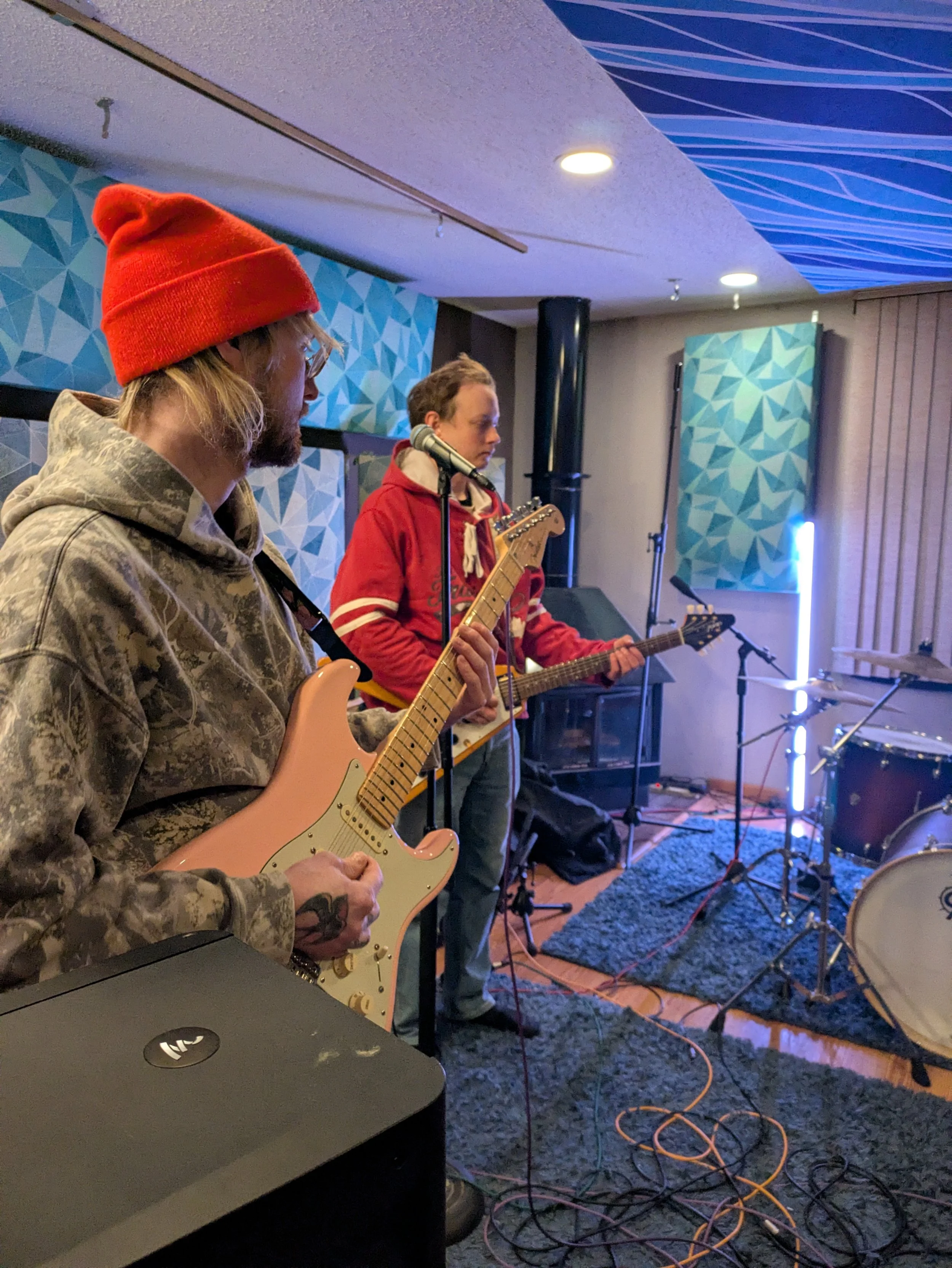 Two musicians are playing guitar, one with a pink electric guitar and the other with an acoustic guitar, in a cozy indoor setting with a textured wall, geometric wall decor, and bass drum, microphone, and various cables on the floor.