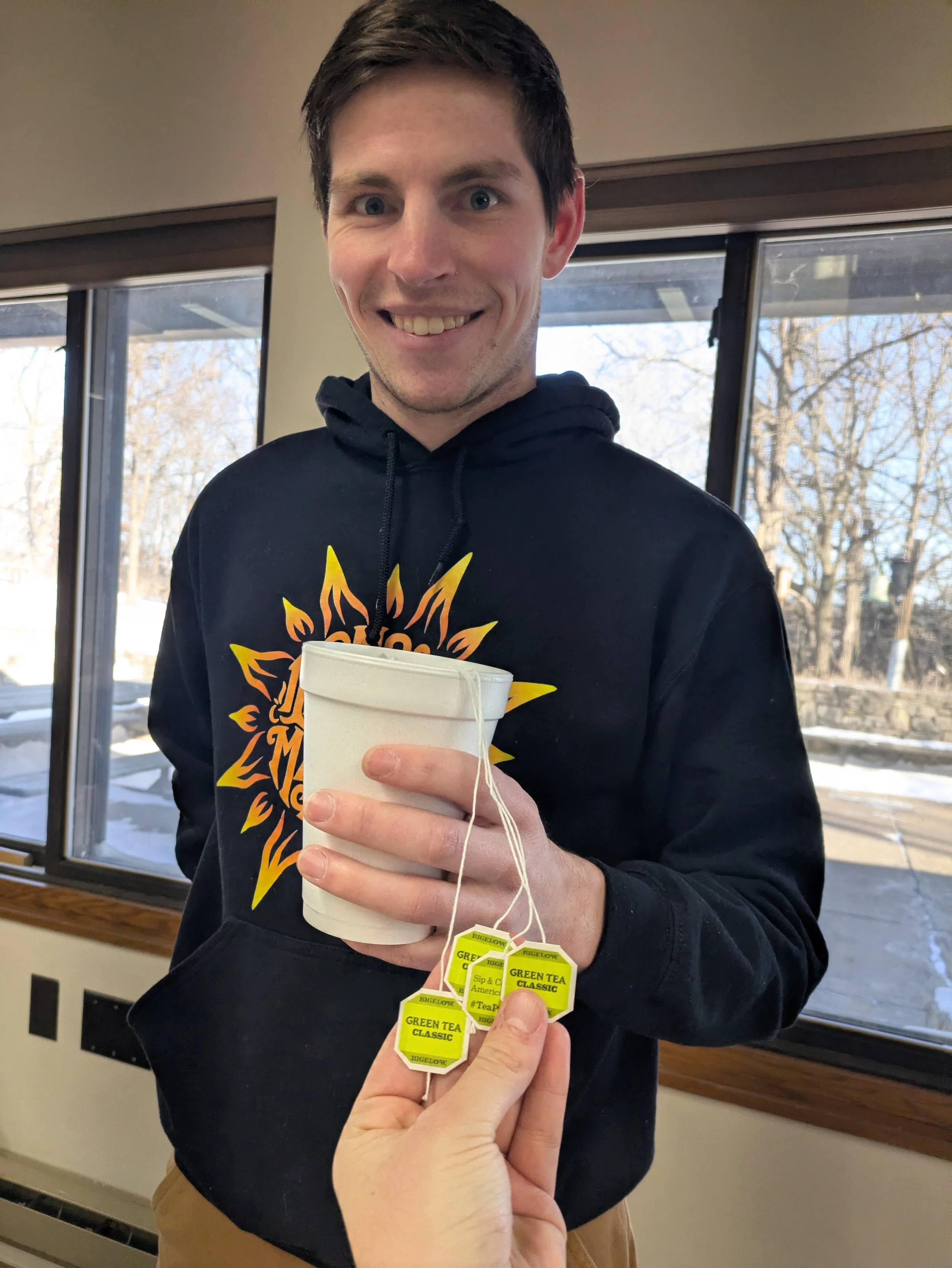 A young man with short dark hair and a black hoodie displaying a graphic, smiling and holding a cup of tea, with a background of windows showing trees outside. A hand in the foreground offers tea tags reading 'GREEN TEA CLASSIC'.