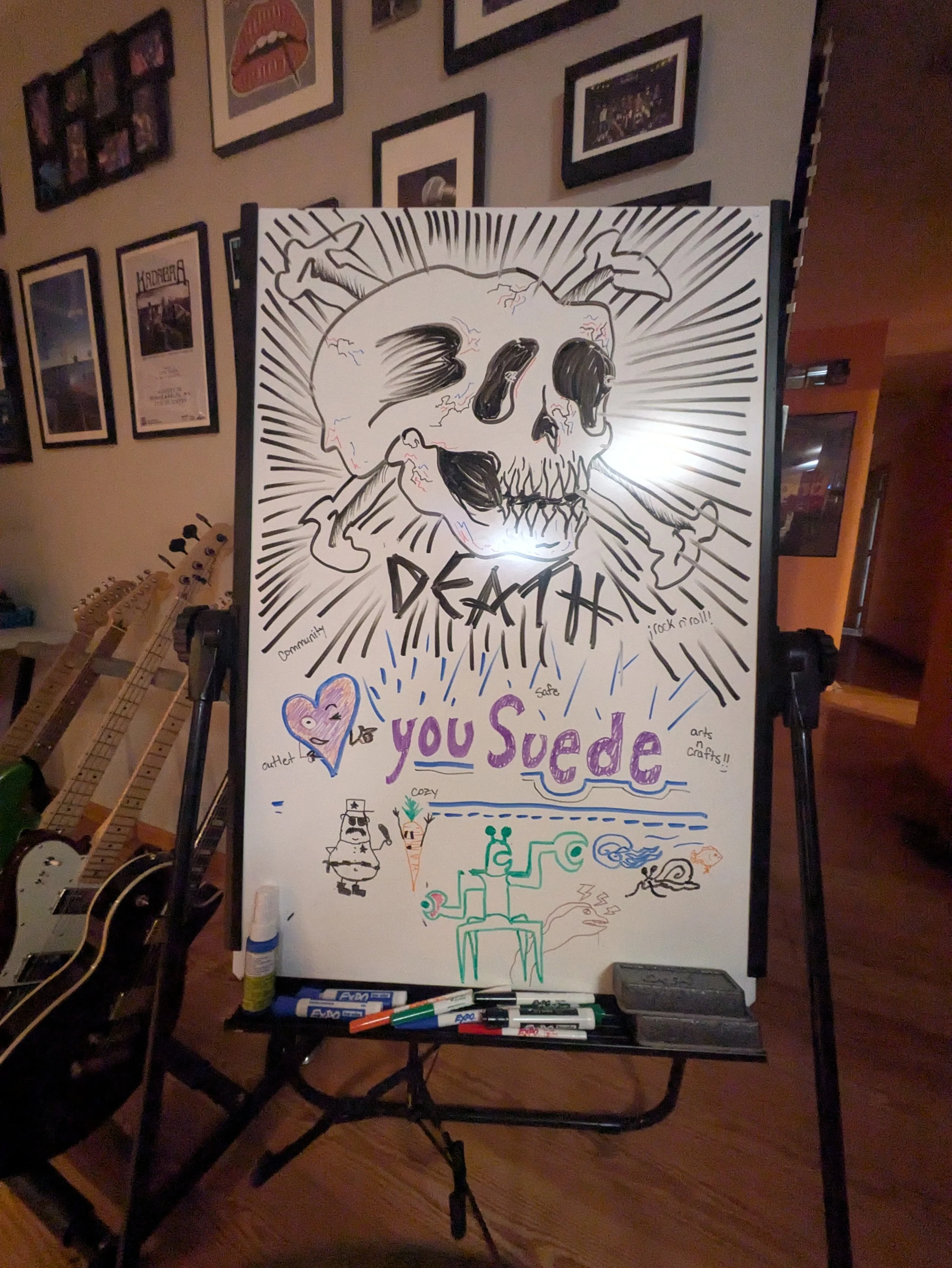 A whiteboard with a large skull illustration surrounded by radiating lines, featuring handwritten words and small drawings, including a purple heart, a cupcake, and a snake, with pens and markers on a tray below and guitar images on the wall.