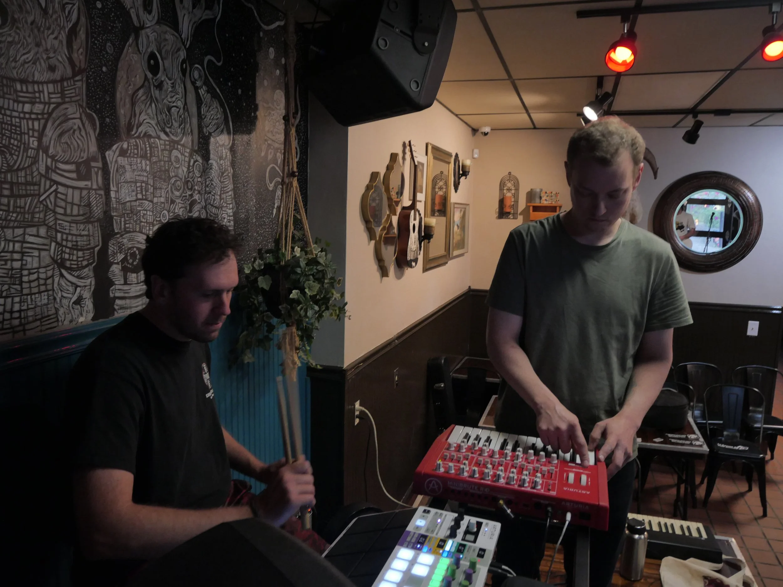 Two men are in a dimly lit room, one playing a keyboard and the other adjusting electronic music equipment. The room is decorated with artwork on the wall, including a black and white mural, framed pictures, and decorative mirrors. Red and white stag