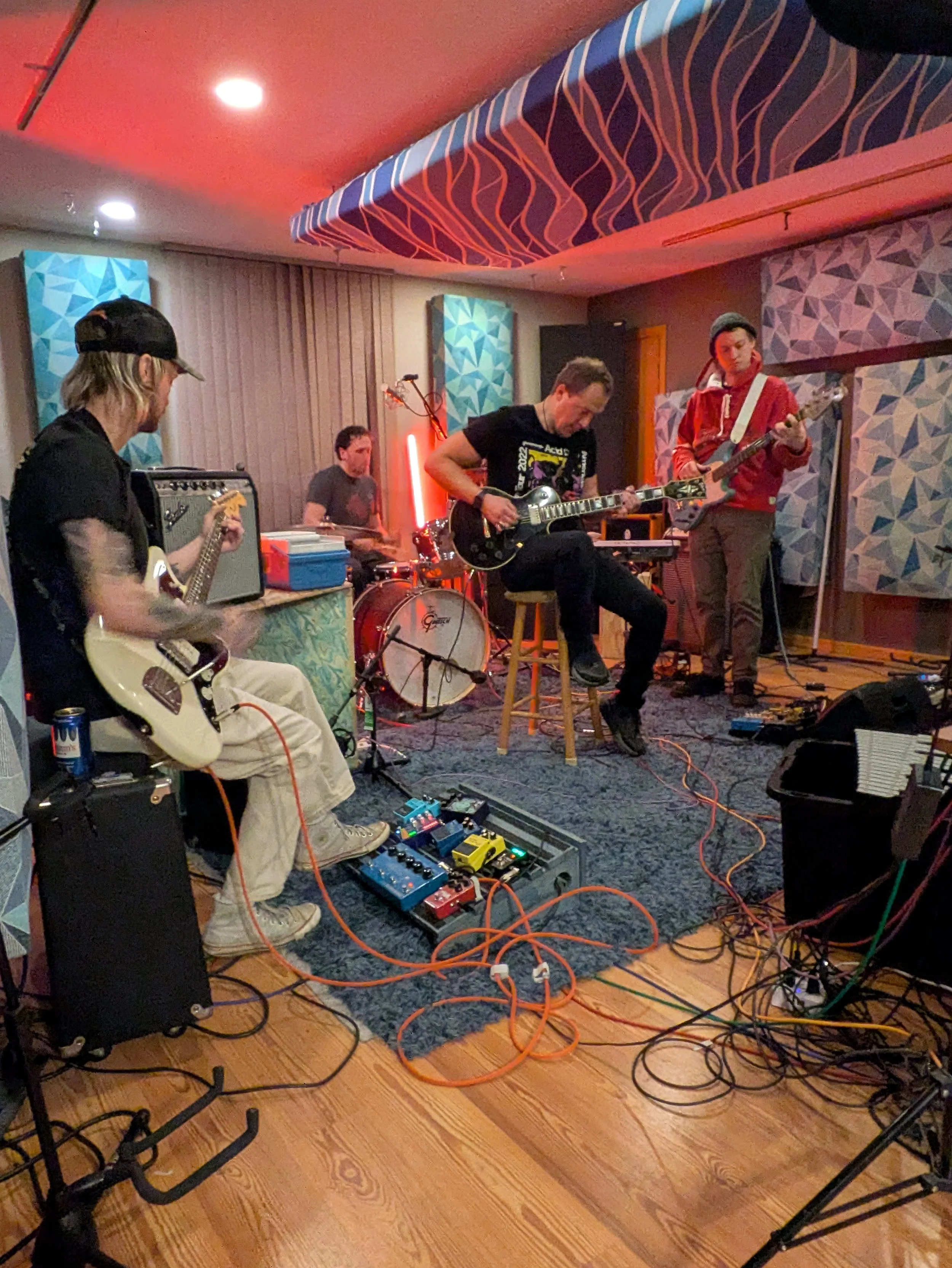 A band of four musicians playing instruments in a room with patterned blue and pink walls, wooden floor, and ceiling with hanging fabric or acoustic panels.