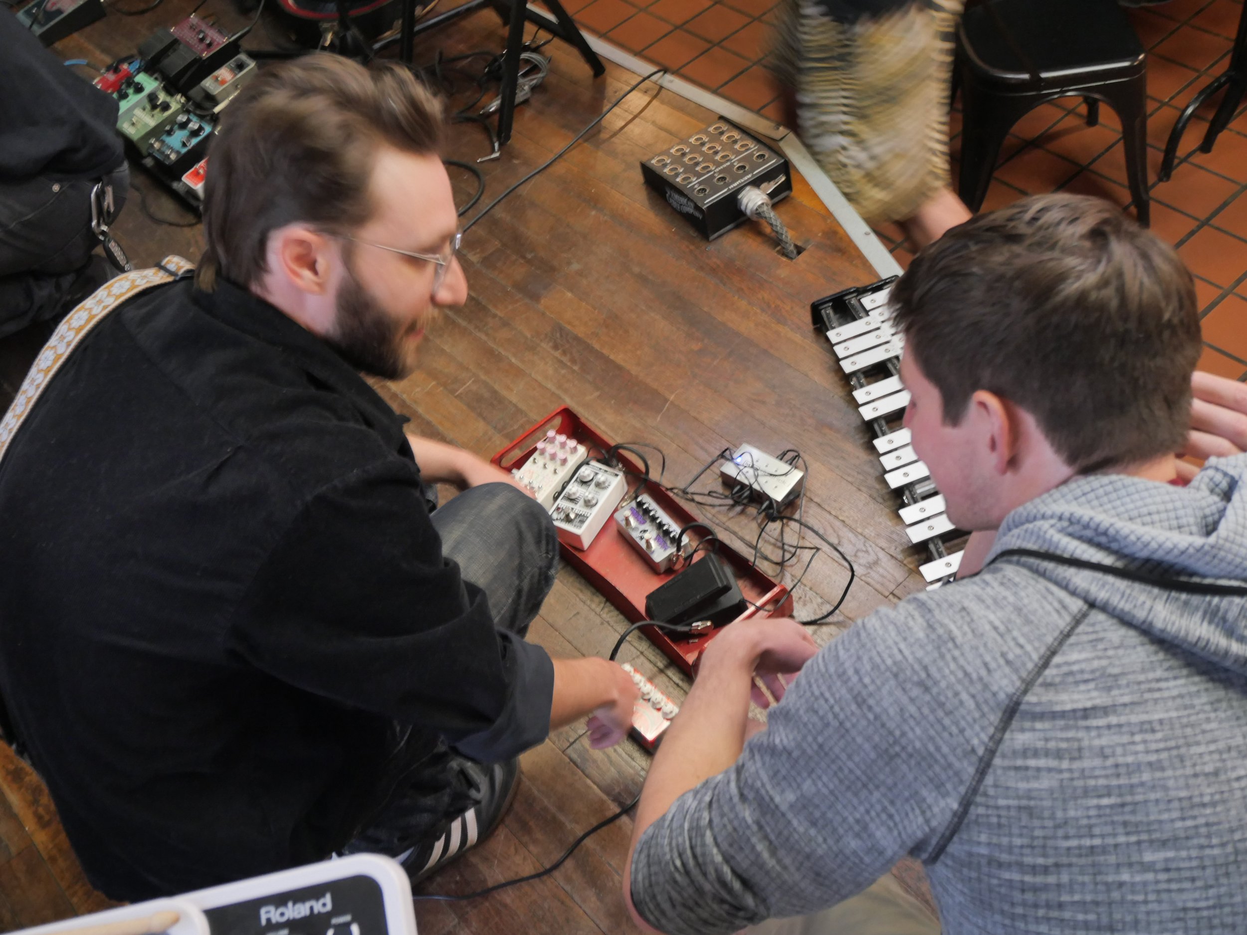 Two men are sitting on the floor playing with electronic music equipment, including a small electronic drum pad and various pedals, with wires and other devices around them, in a room with tiled flooring.