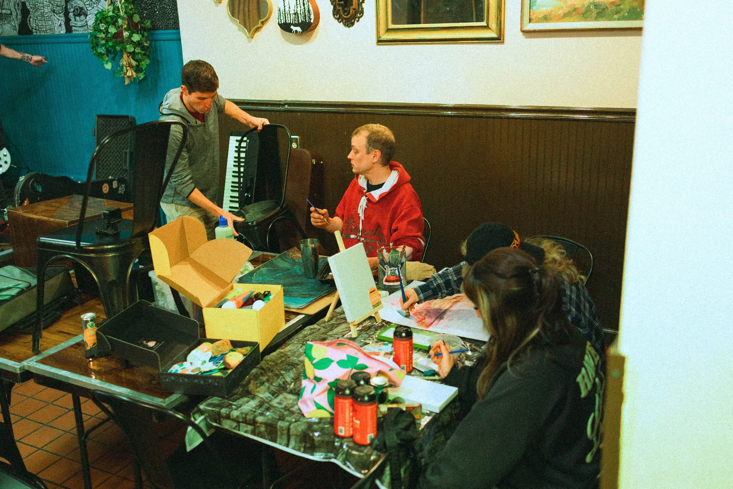 People gathered around a table painting and doing arts and crafts in a room decorated with framed artwork and wall hangings.