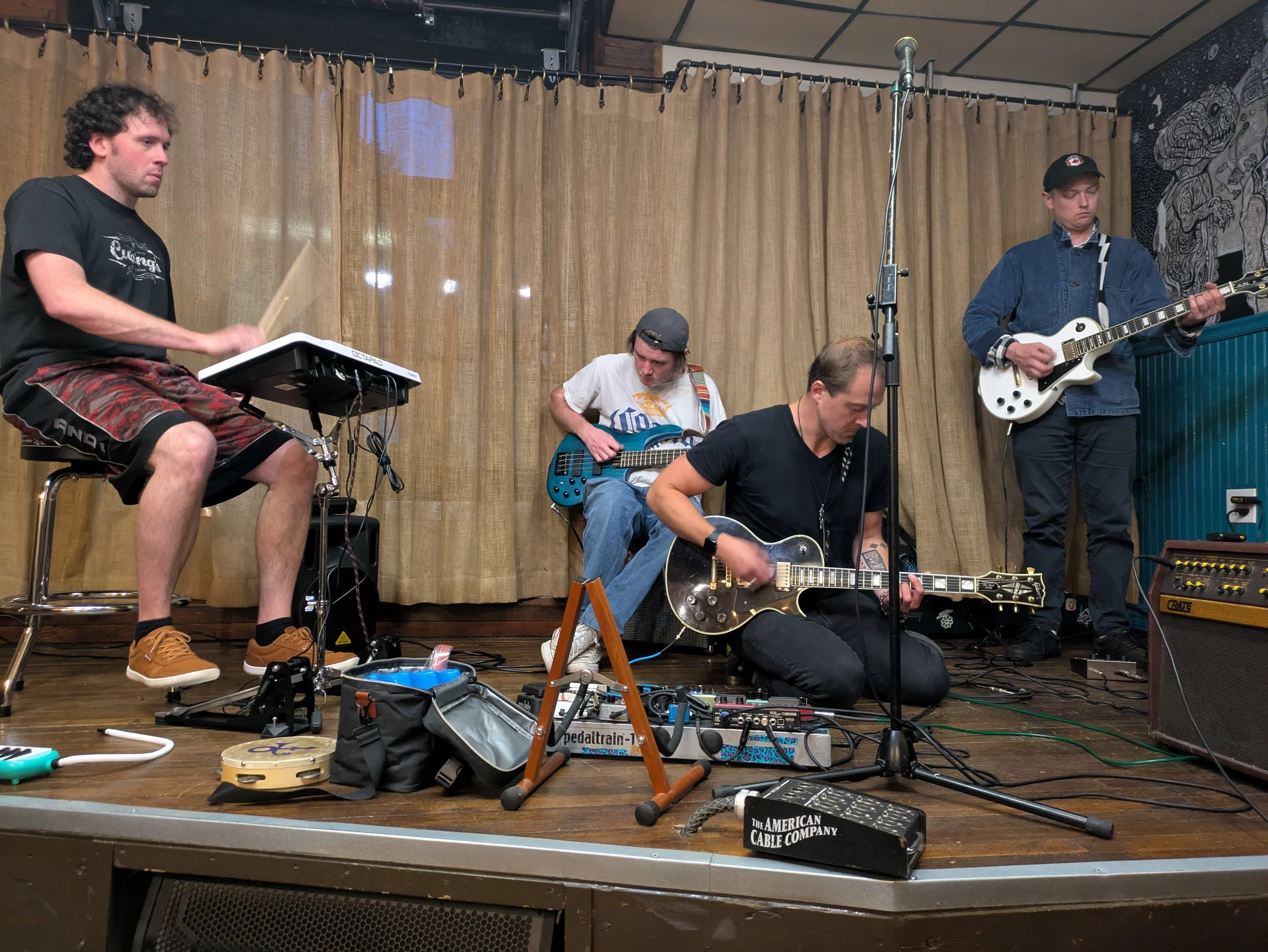 A group of four musicians performing on a stage with musical instruments and equipment. One person is playing drums, one is playing bass guitar, another is sitting with an electric guitar, and the fourth is playing an electric guitar. The stage has a