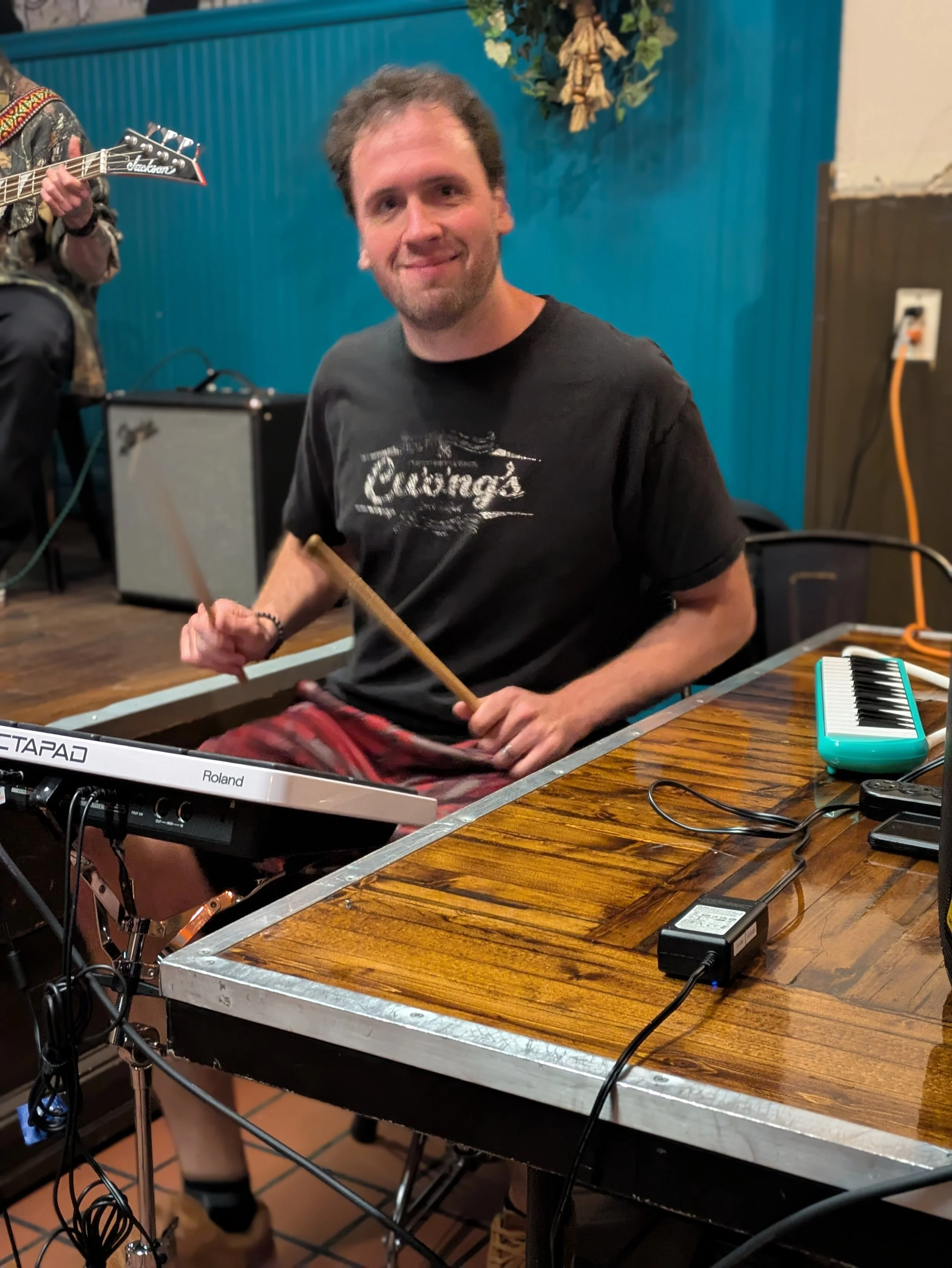A man sitting at a wooden table with musical equipment, including a keyboard, electric guitar, and cables, in a room with blue walls and tan tile floors.