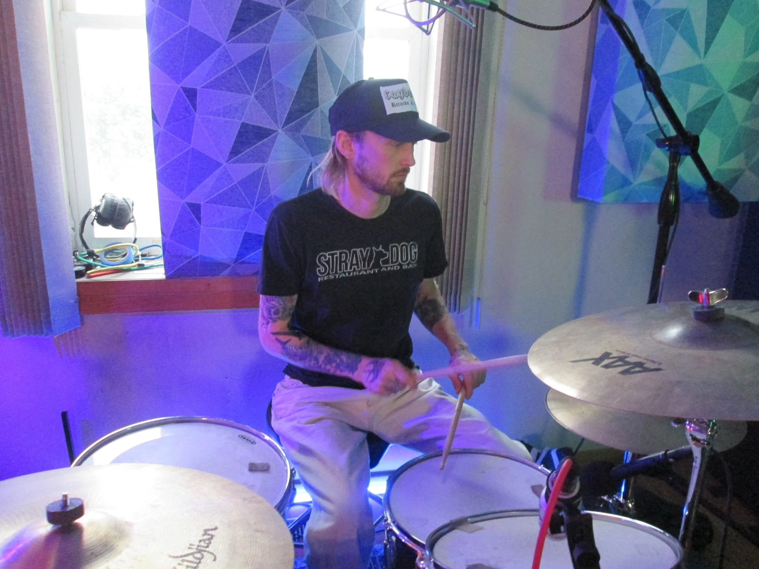 A man playing drums in a room with colorful geometric patterned wall art, wearing a black cap and T-shirt, with a microphone overhead.