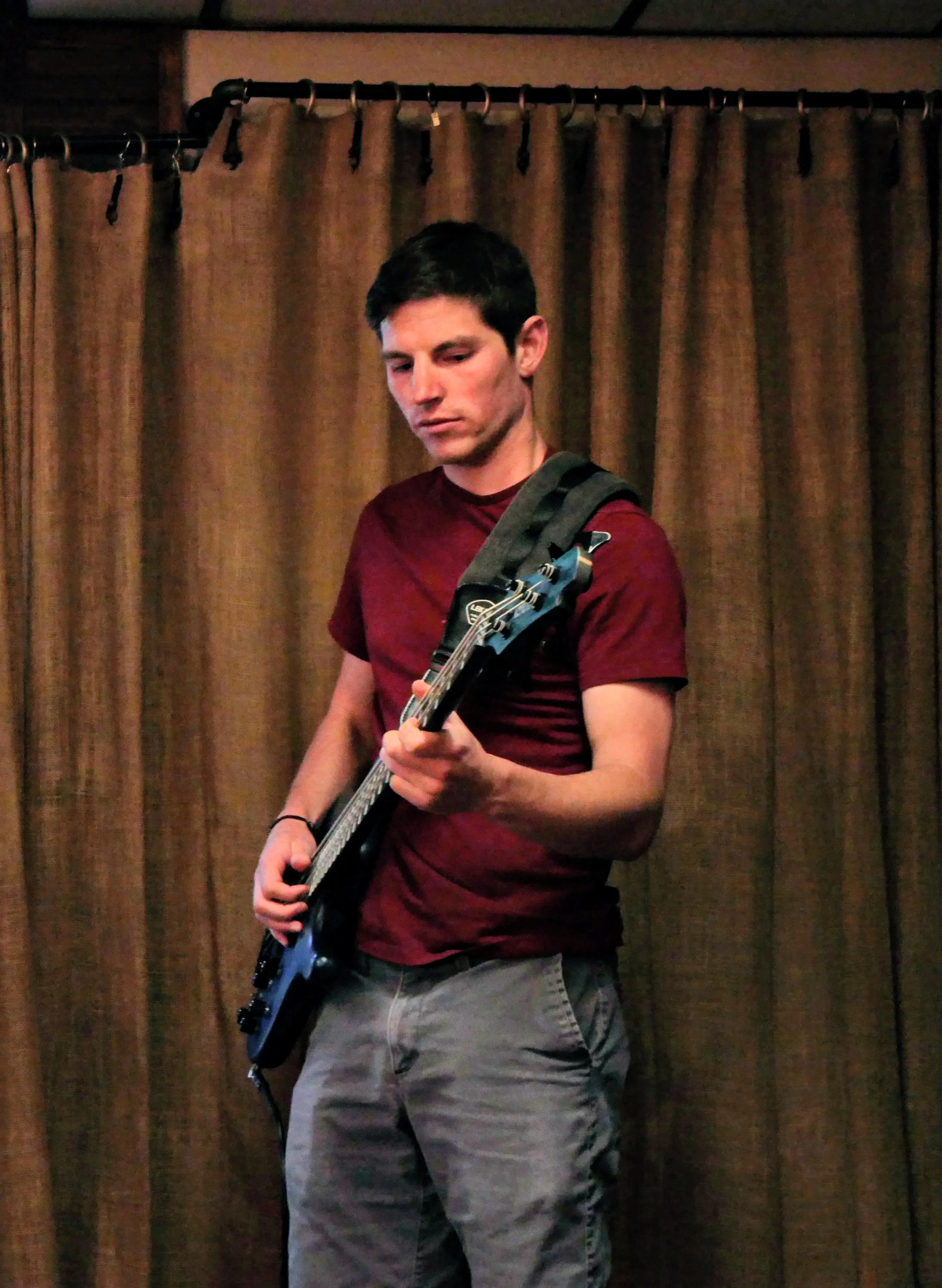 A young man with short dark hair playing an electric guitar in front of brown curtains.