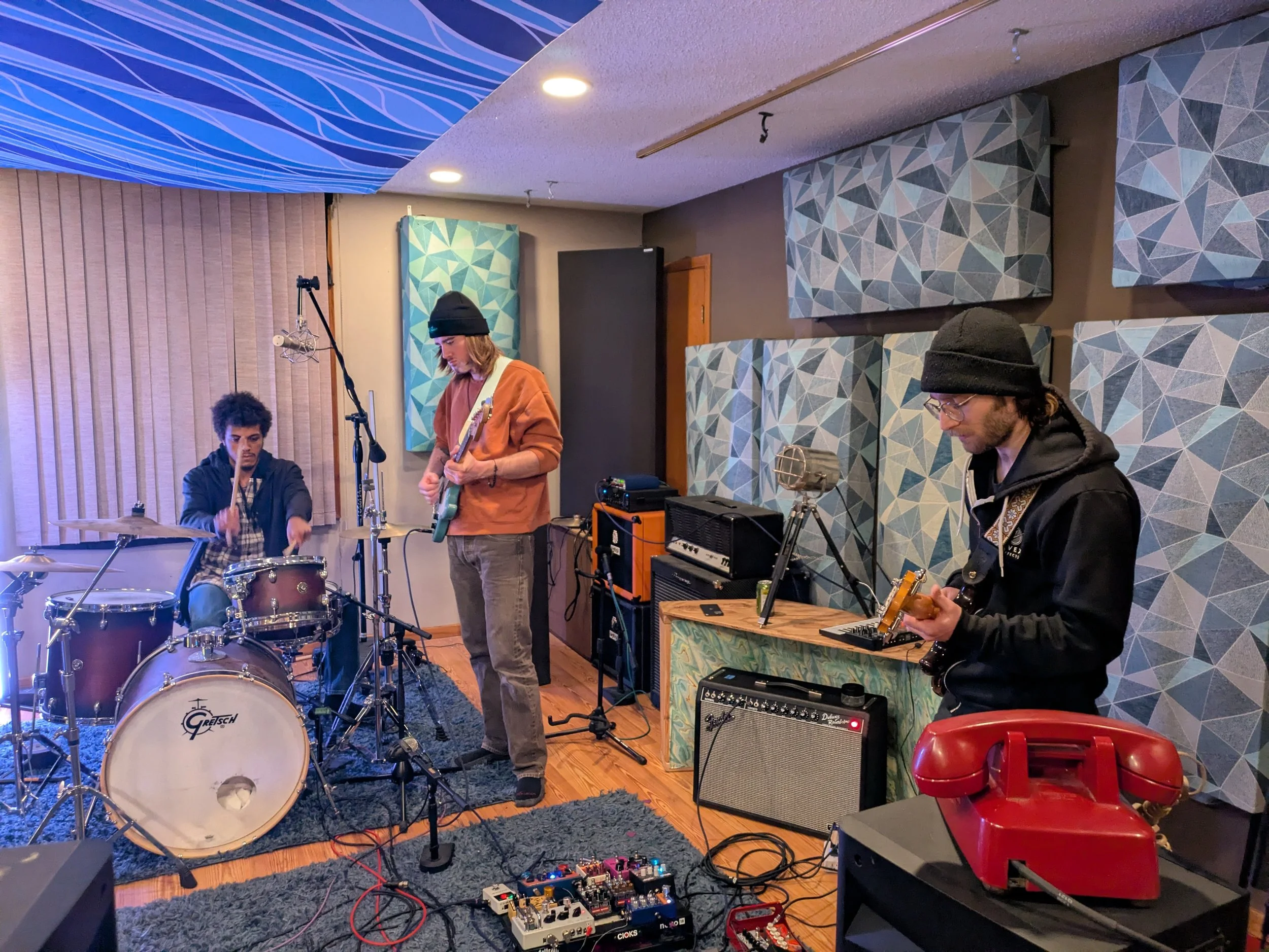 Four people are playing musical instruments in a recording studio. One person is sitting at a drum set, another is standing with a guitar, and two others are playing electric guitar. The room has soundproof panels on the walls, and there are various 