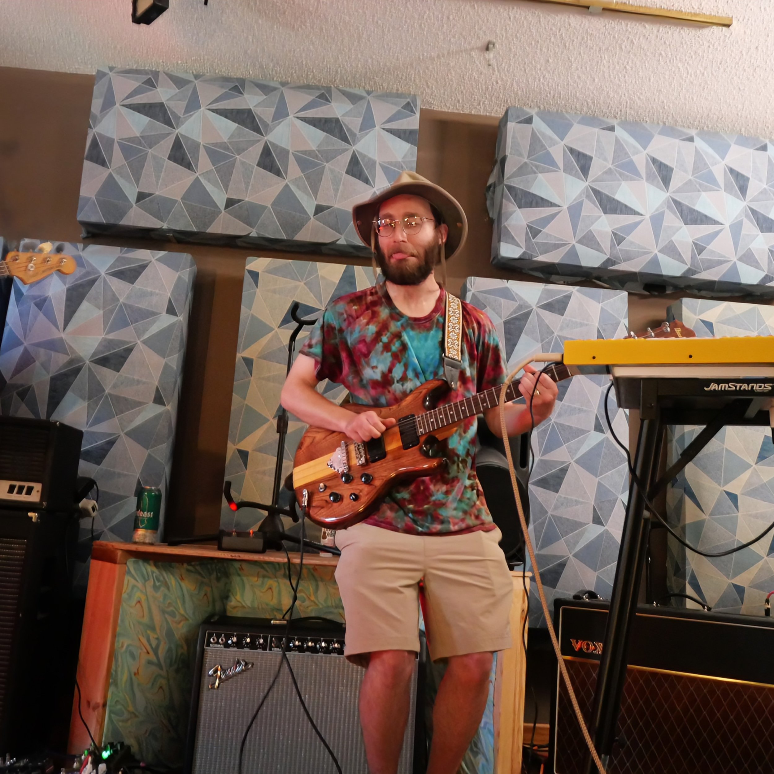 A man with glasses and facial hair wearing a bucket hat and colorful tie-dye shirt playing an electric guitar in a music room with patterned wall panels, amplifiers, and musical equipment.