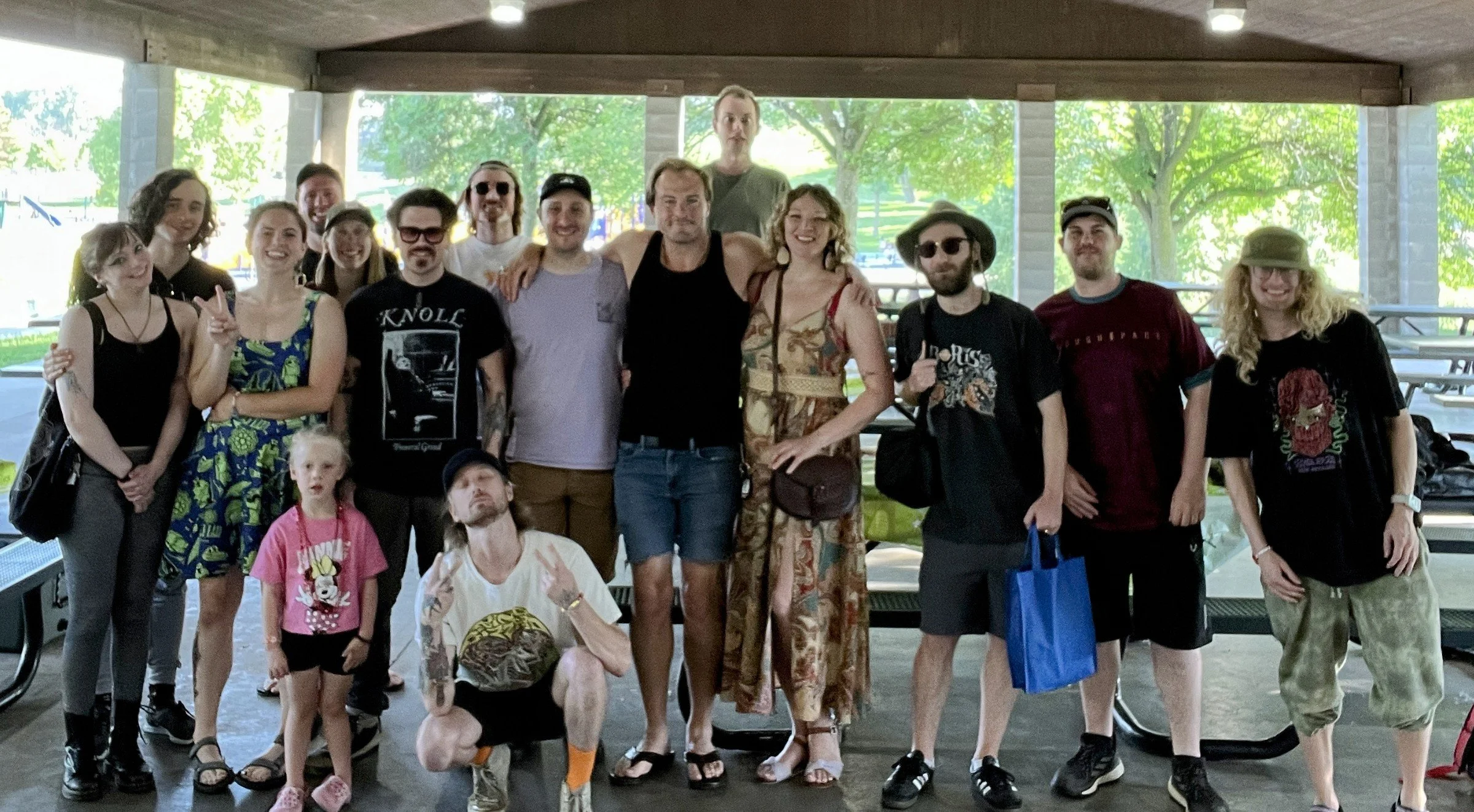 A group of diverse people, including children and adults, posing together outdoors under a shelter with green trees in the background. Some are smiling and making peace signs.