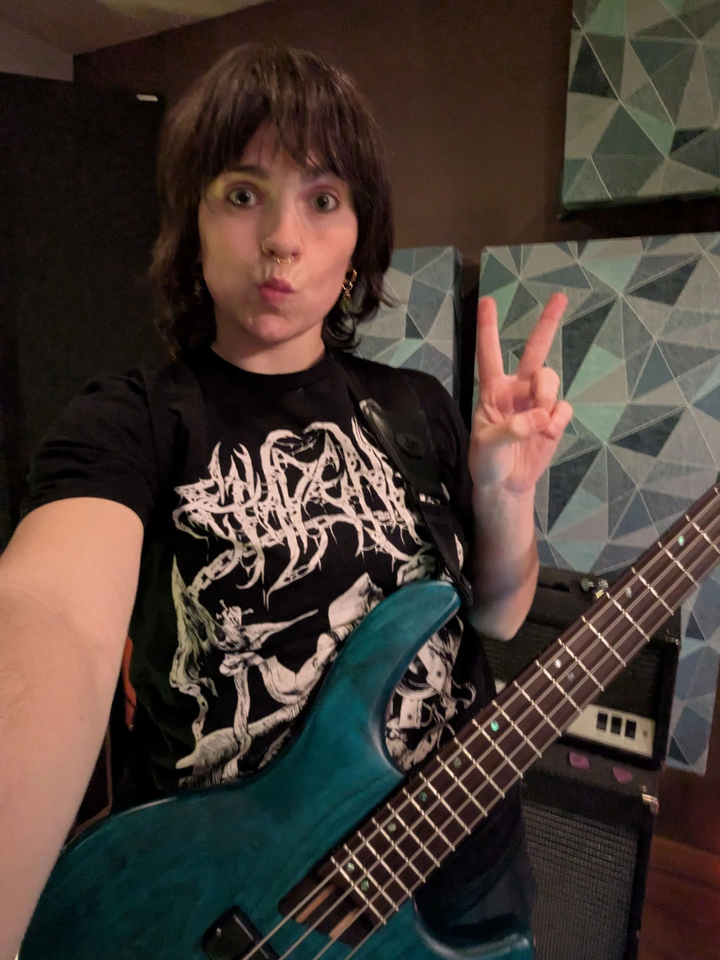A person with dark hair making a peace sign while holding an electric guitar. The person is indoors, with a geometric-patterned wall and guitar amplifier visible in the background.