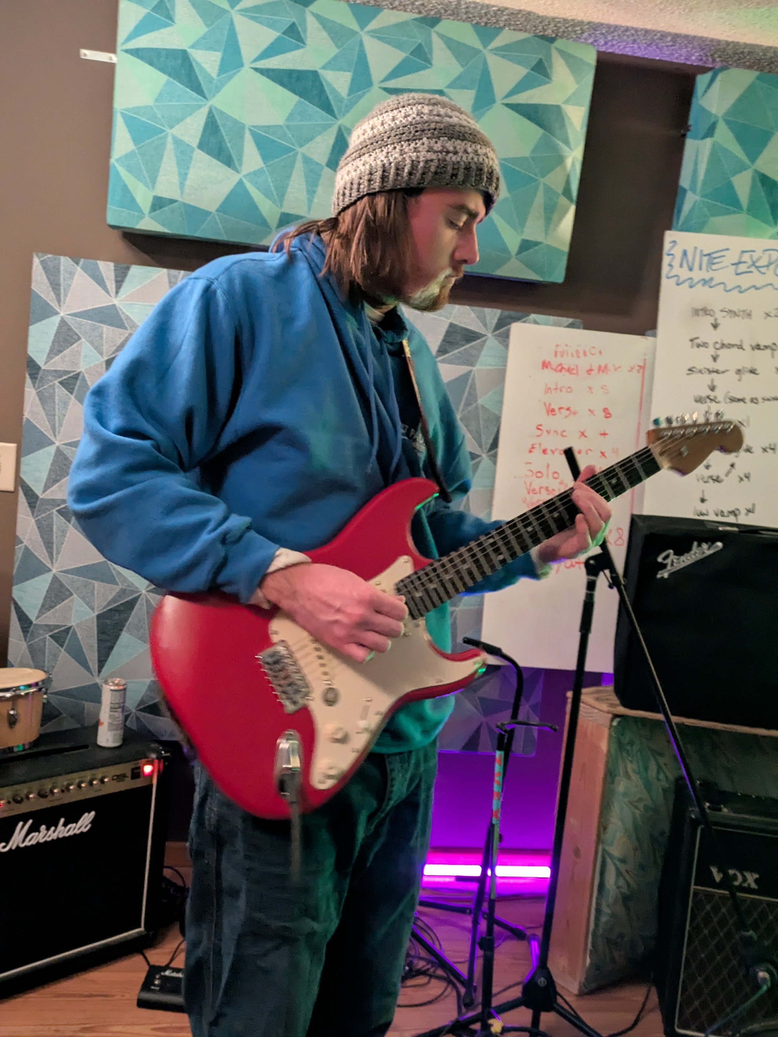 A man with shoulder-length hair wearing a gray knit hat and a blue jacket playing a red electric guitar in a music rehearsal space with amplifiers and whiteboards with musical notes and song lyrics.
