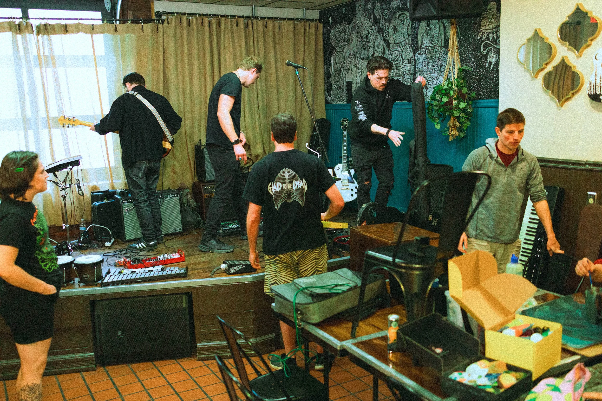 A group of people preparing for a live music performance in a cozy room. Some are tuning instruments, others are setting up equipment. The room has warm lighting, wall art, and a table with various supplies.