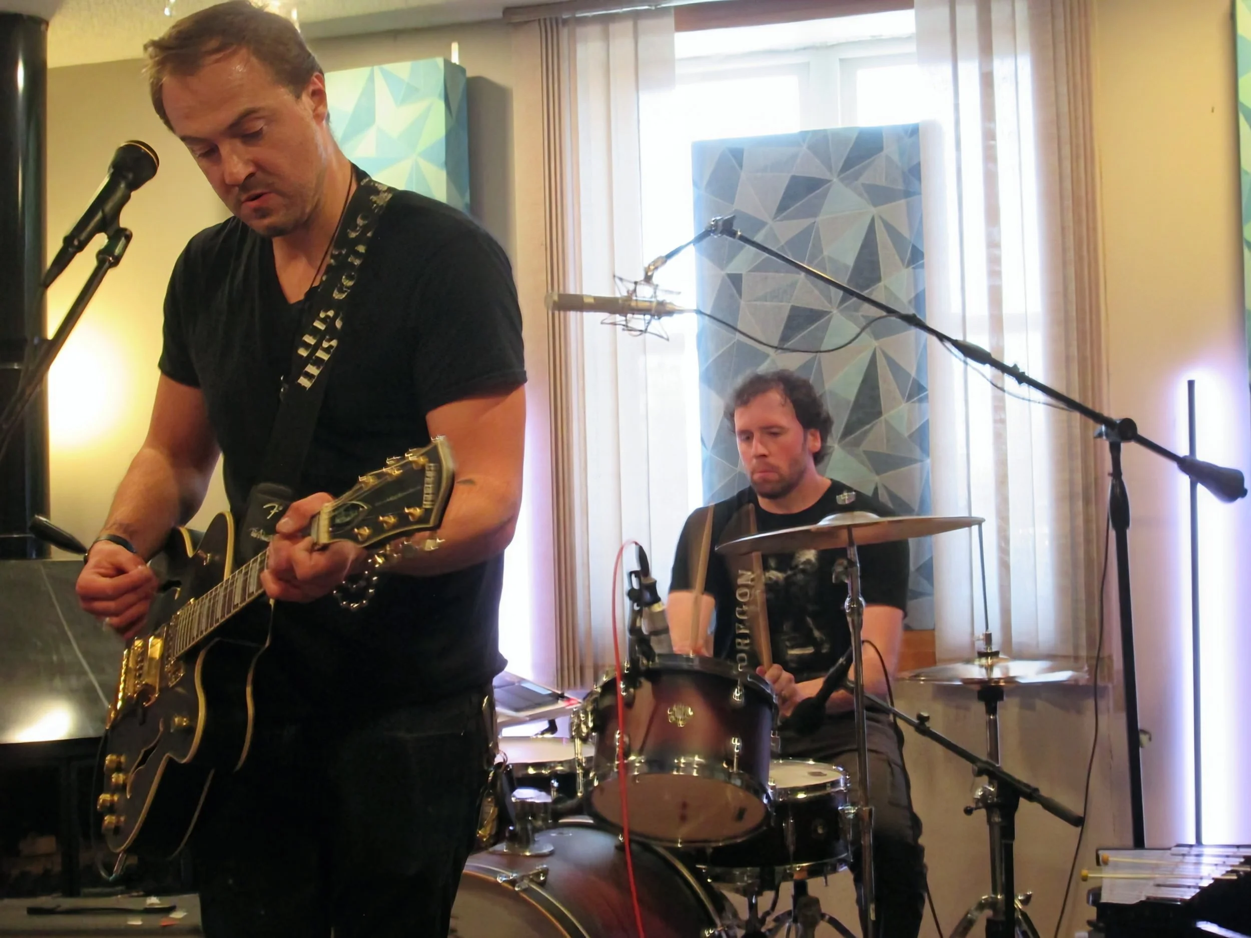 Two musicians performing indoors, one playing an electric guitar and the other playing a drum set, in front of large windows with natural light.