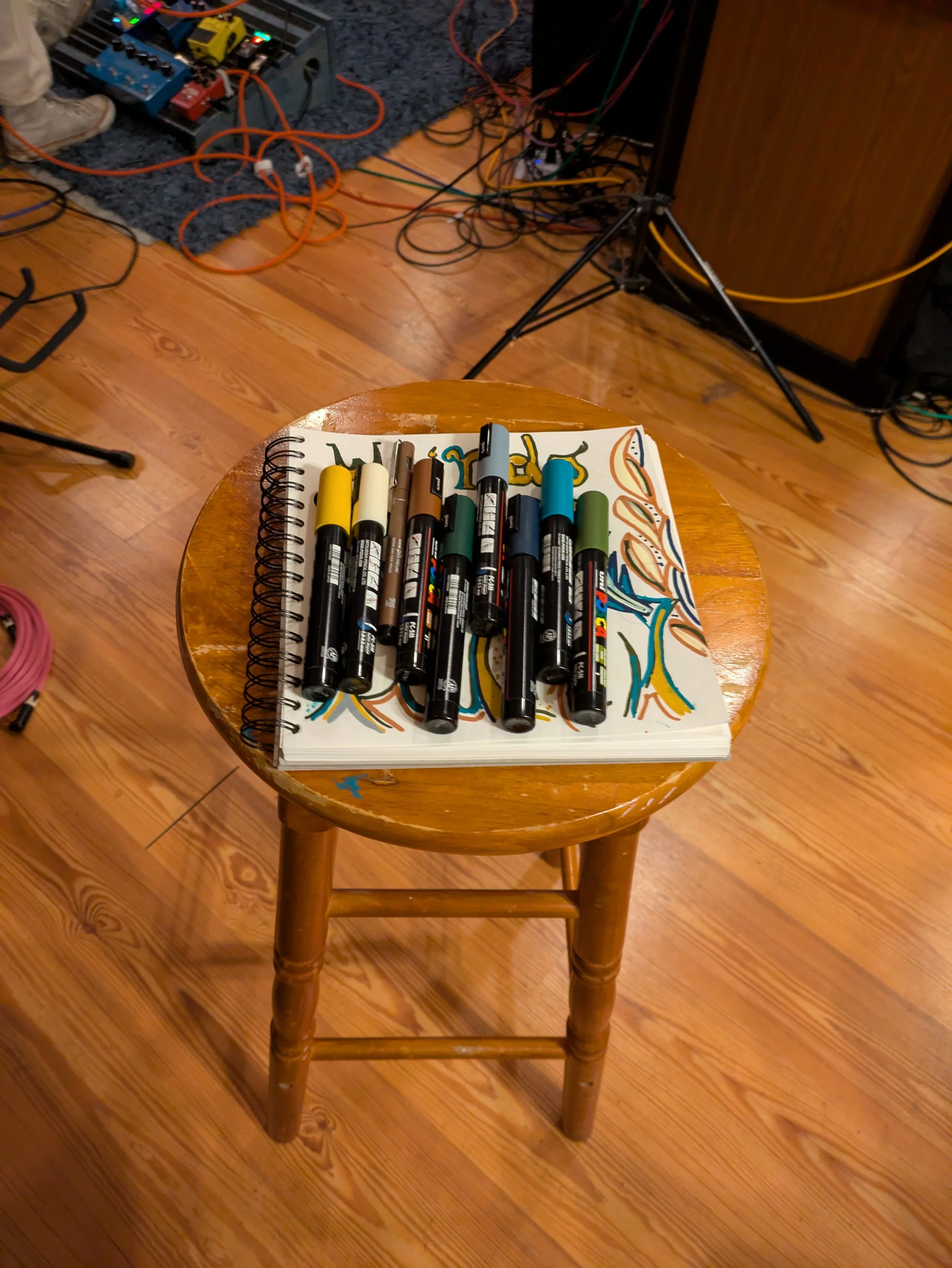 A wooden stool with a spiral notebook on top, covered with colorful markers. In the background, there is a sound equipment setup with cables and effects pedals on a carpeted floor.