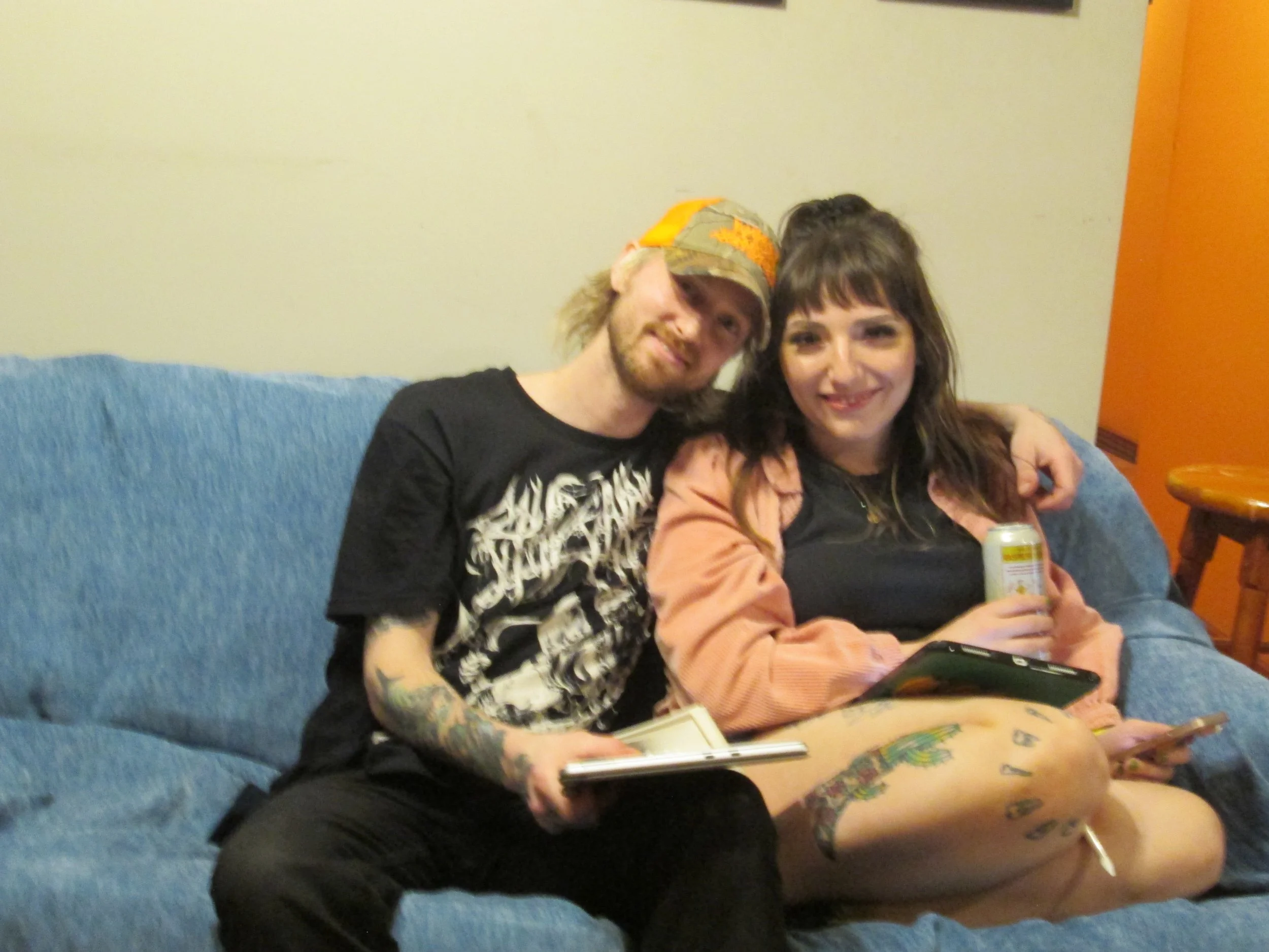 Two friends sit together on a blue couch, smiling at the camera. The person on the left has long blonde hair, tattoos, and is wearing a black T-shirt, while the person on the right has shoulder-length brown hair, and is wearing a black shirt and an o