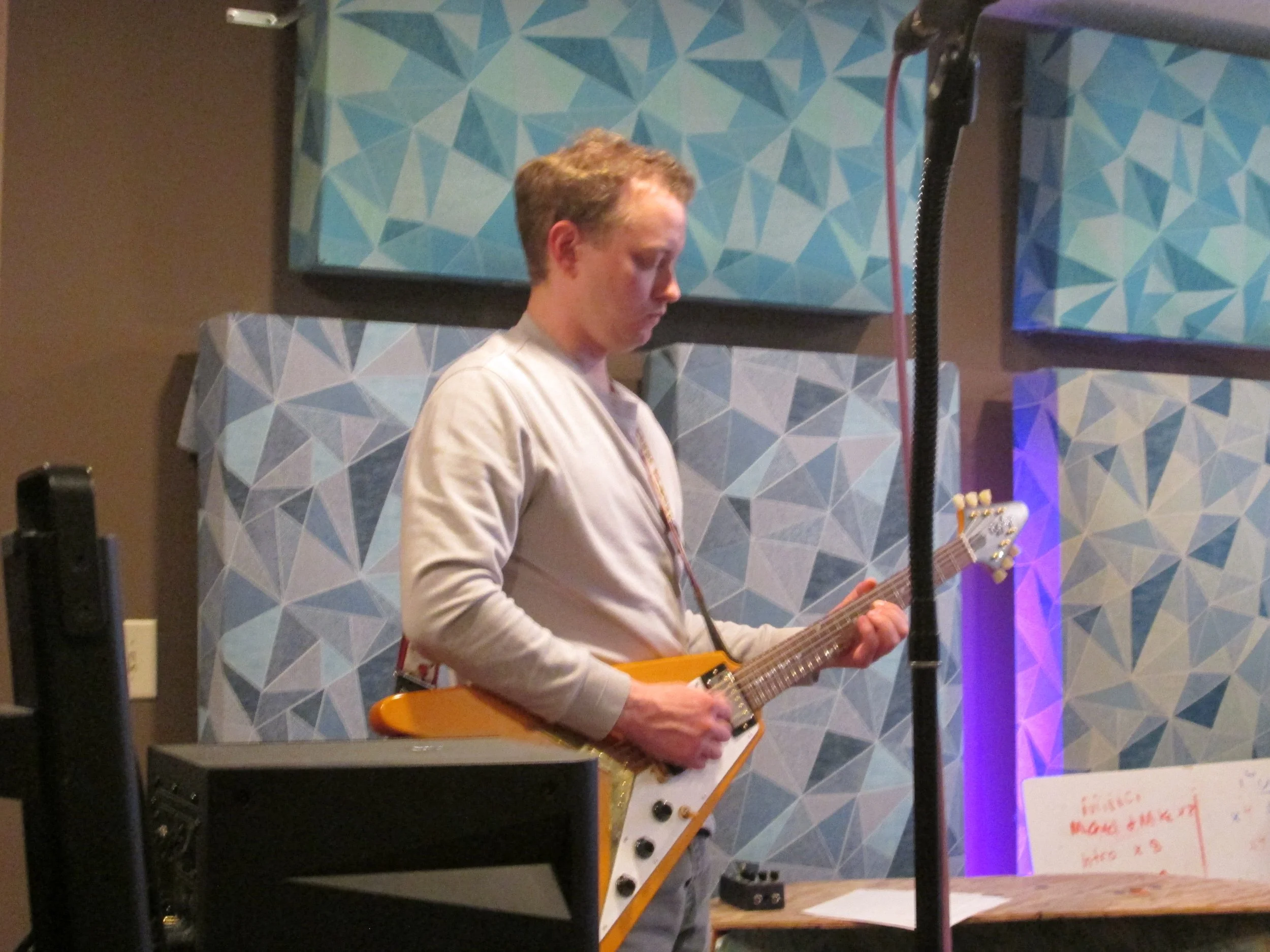 A person with short, curly hair playing an electric guitar on stage against a decorative blue geometric patterned backdrop.
