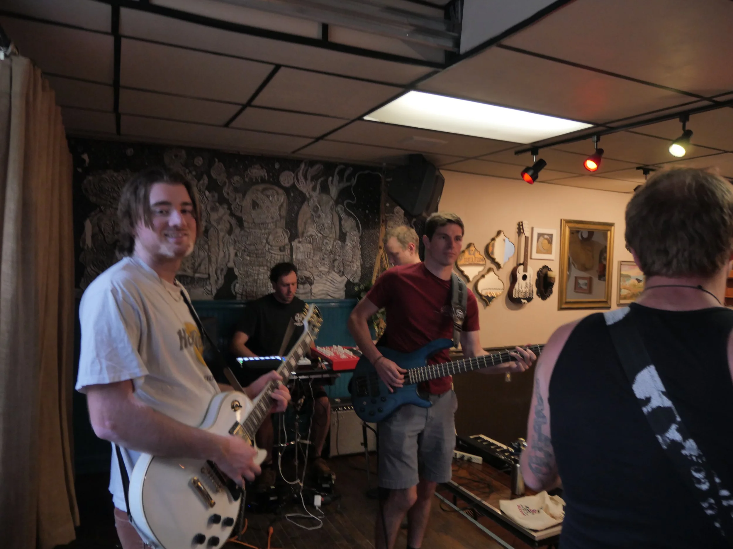 Group of people playing musical instruments, such as guitars, in an indoor space decorated with framed pictures and a painting on the wall.