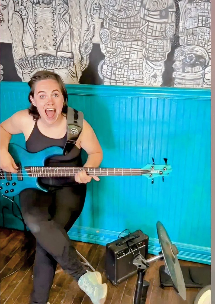 A woman playing a blue guitar in a room with teal paneling and black and white artwork on the wall, looking excited or happy.