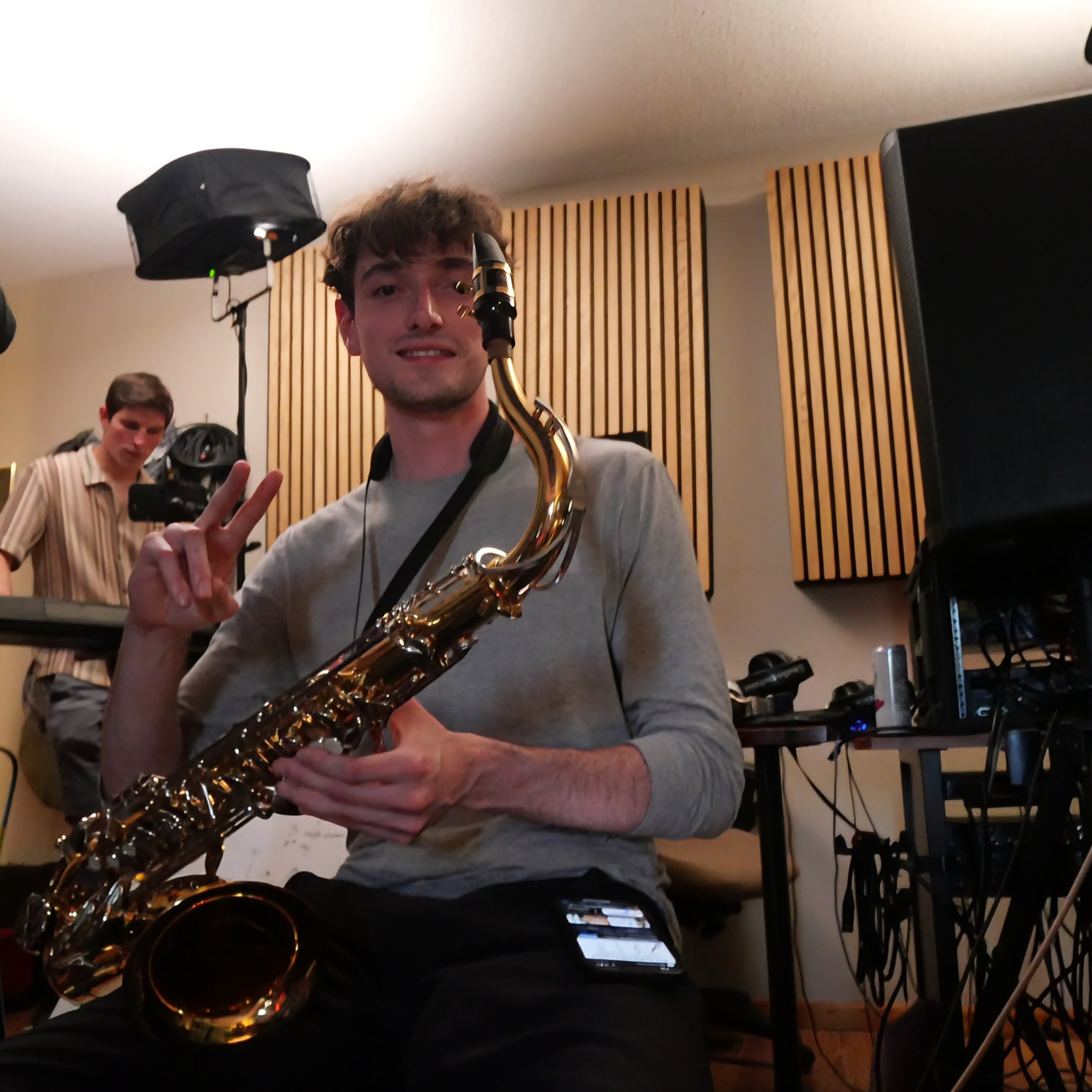 A young man holding a saxophone and making a peace sign with his fingers, in a room with acoustic wood wall panels, other people in the background, and music equipment.